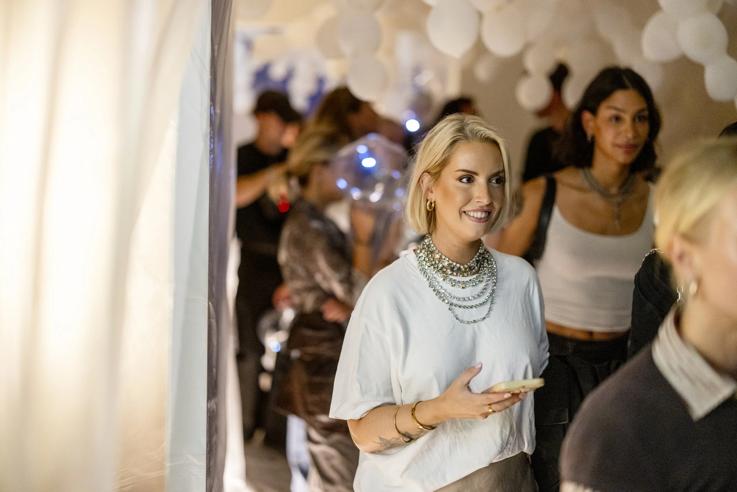 A woman with short blonde hair, wearing a white shirt with multiple necklaces, holding a phone, smiling at an indoor event.