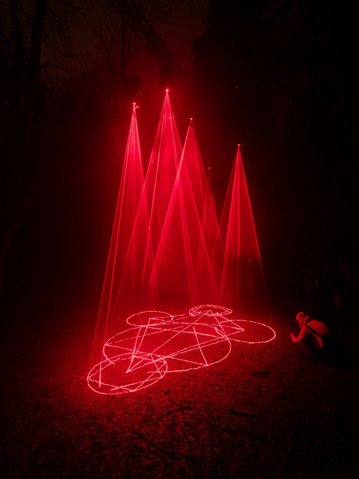 Cathedral of Ego: Light, Code, and Transcendence  MAXIME PLESCIA-BÜSCHI AND BJØRN STAAL  x COLLECTIVE OSLO A person in a dark outdoor setting with red laser lights creating geometric shapes and cone-like structures in the air.