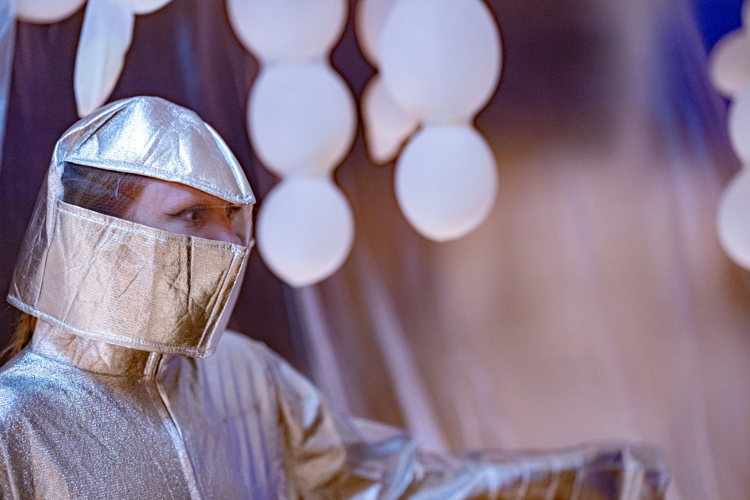 A person wearing a shiny silver protective suit and helmet, with only their eyes visible, in a setting with white circular lights in the background.