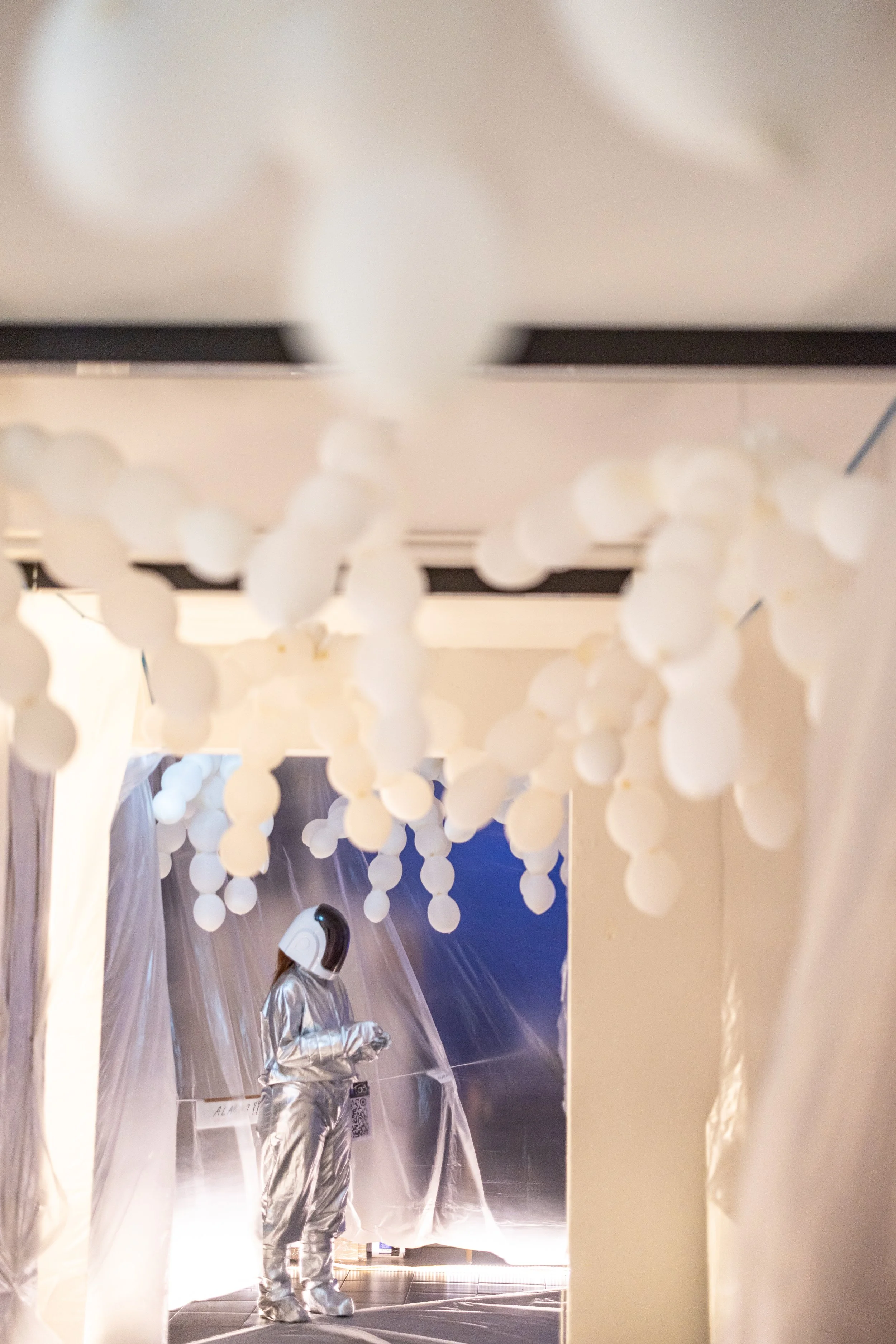 Person in a silver spacesuit standing under hanging white spherical decorations in a futuristic, metallic setting.