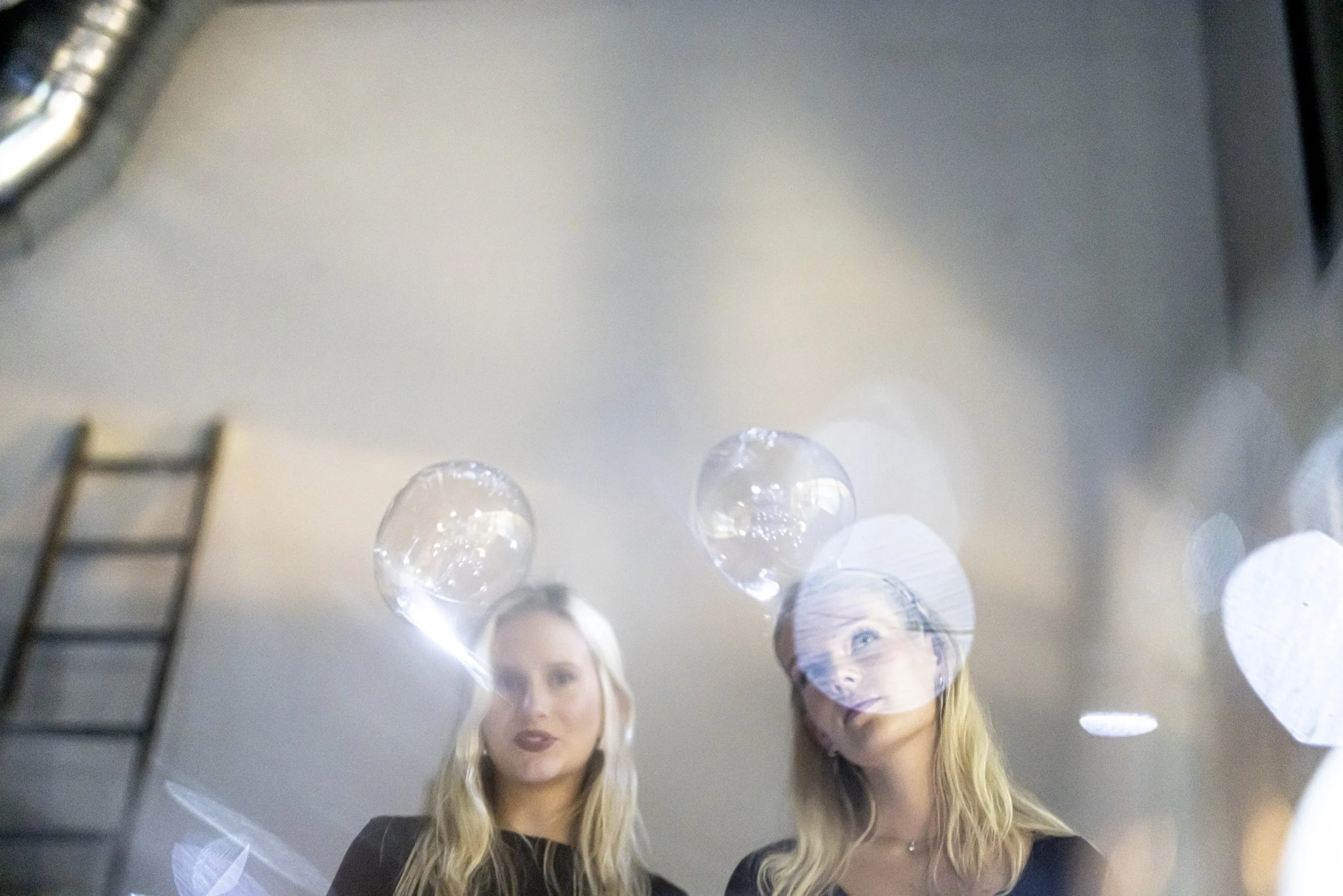 Two women looking into a mirror reflecting bubbles and ceiling lights