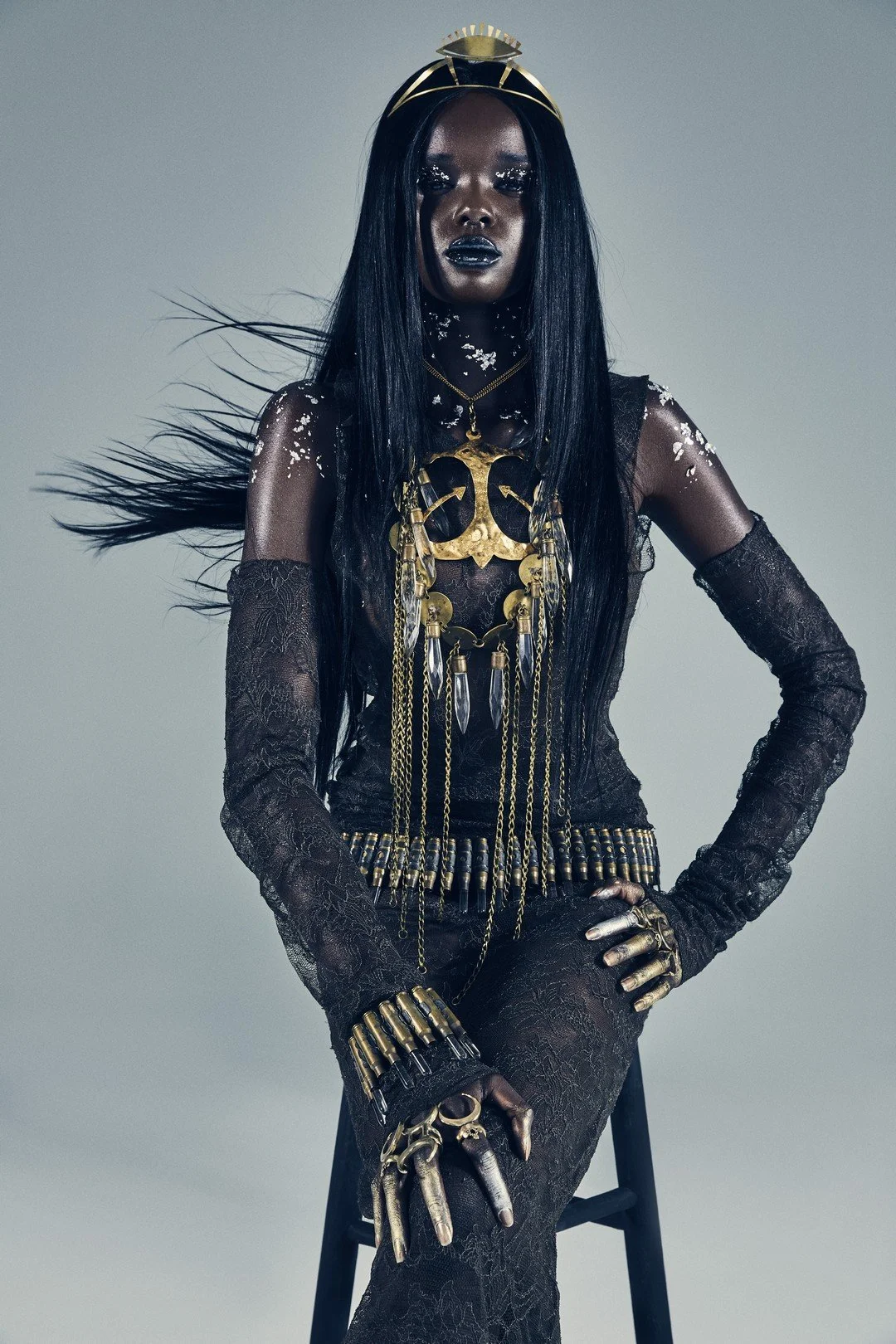 A woman with long black hair and dark makeup, dressed in black lace clothing and adorned with gold jewelry and bone-like accessories, sitting on a black stool against a plain background.