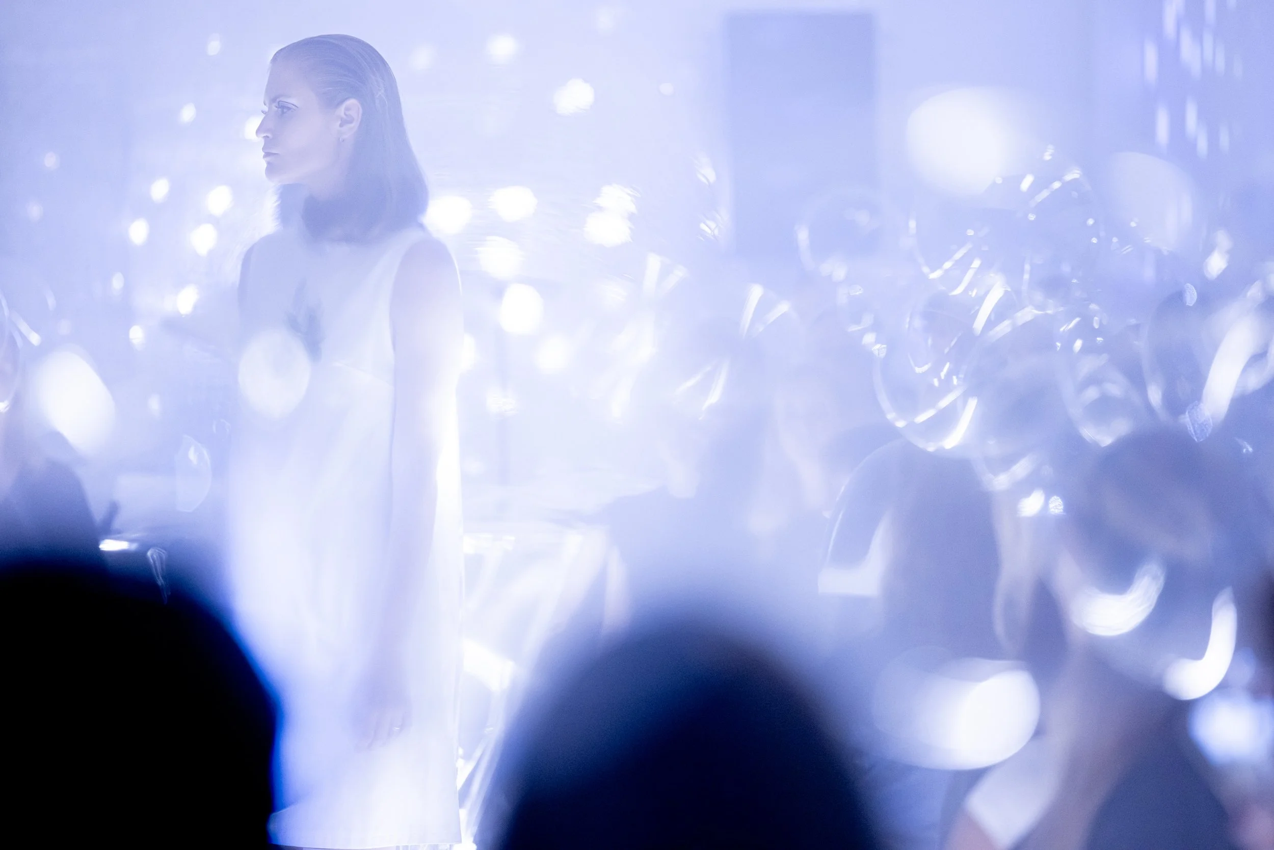 A woman with shoulder-length hair in a white dress standing in a brightly lit, foggy environment with reflective surfaces and blurred silhouettes of people and shiny objects in the background.