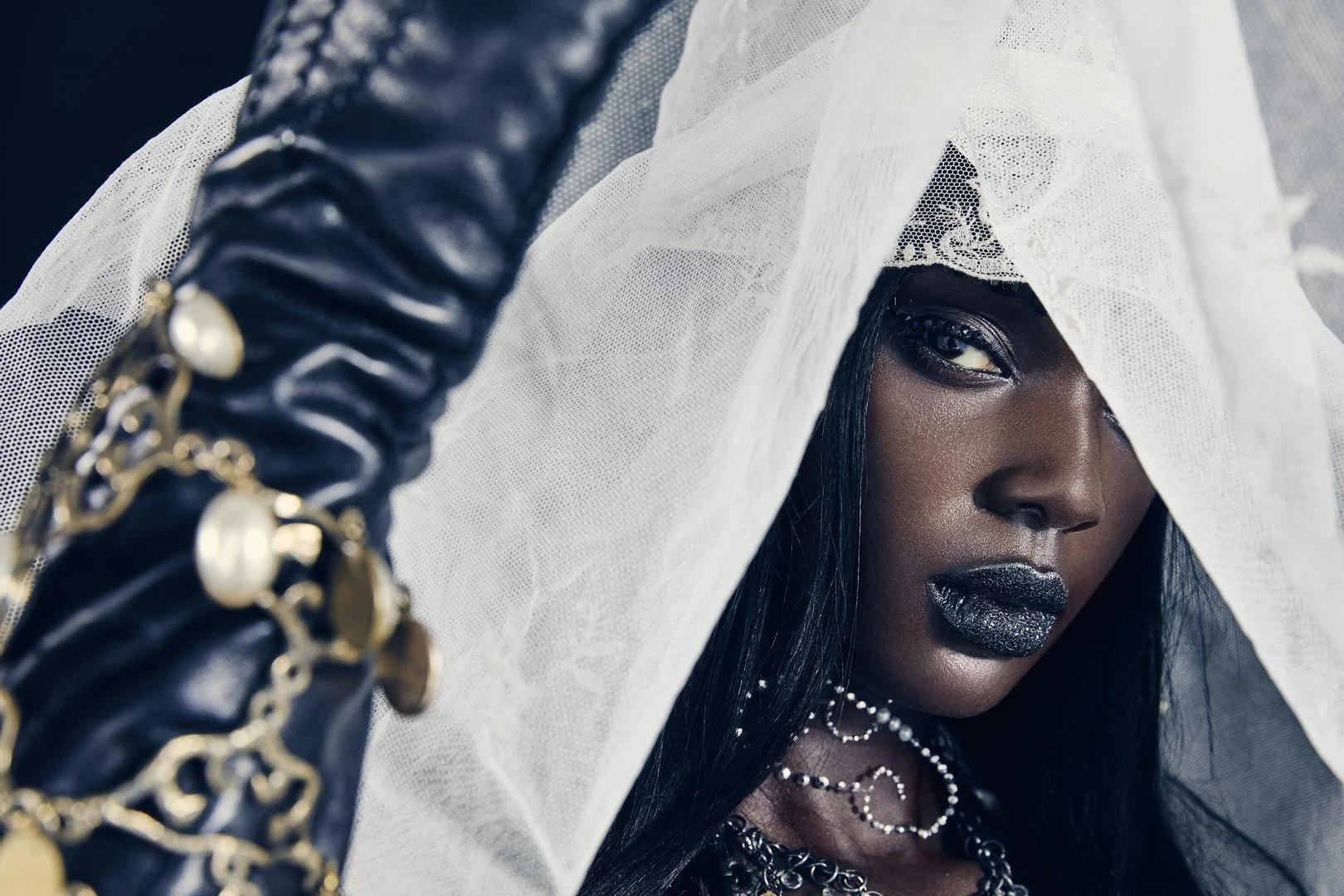 A woman with dark skin and black hair, partially covered by a white veil, wearing black lipstick and jewelry, holding a black leather glove with gold chain accents.