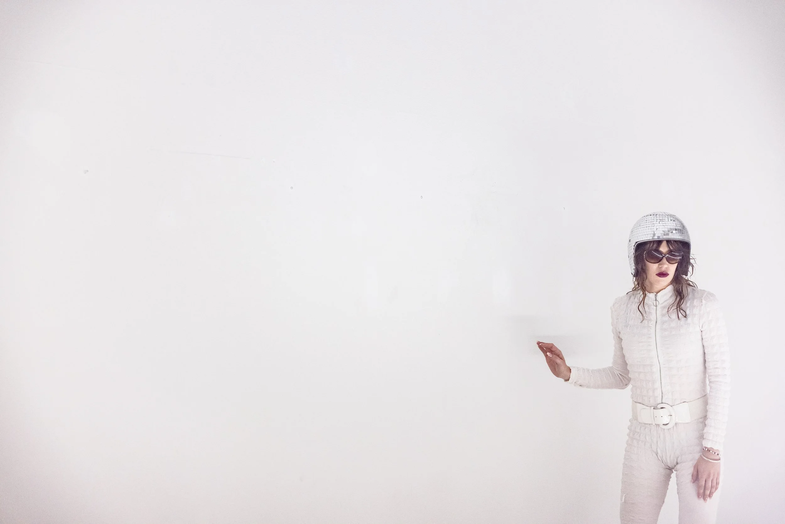 Woman wearing a white textured jumpsuit, large black sunglasses, a shiny silver helmet, and dark lipstick, standing against a white background with her hand raised.