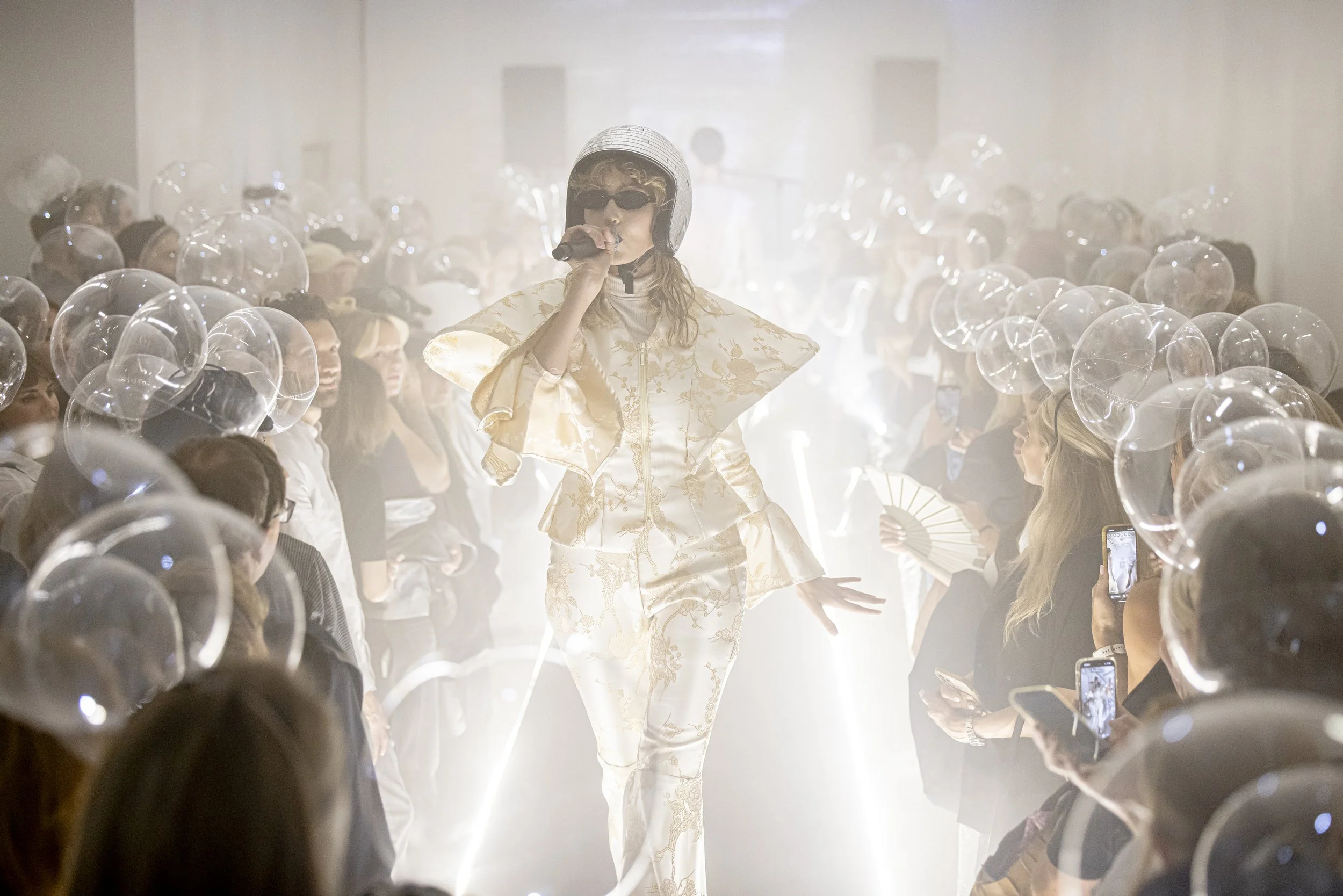 A person walking down a runway during a fashion show, dressed in a shiny, gold-patterned outfit with a wide collar and a helmet, holding a microphone, with an audience on both sides, many taking photos.