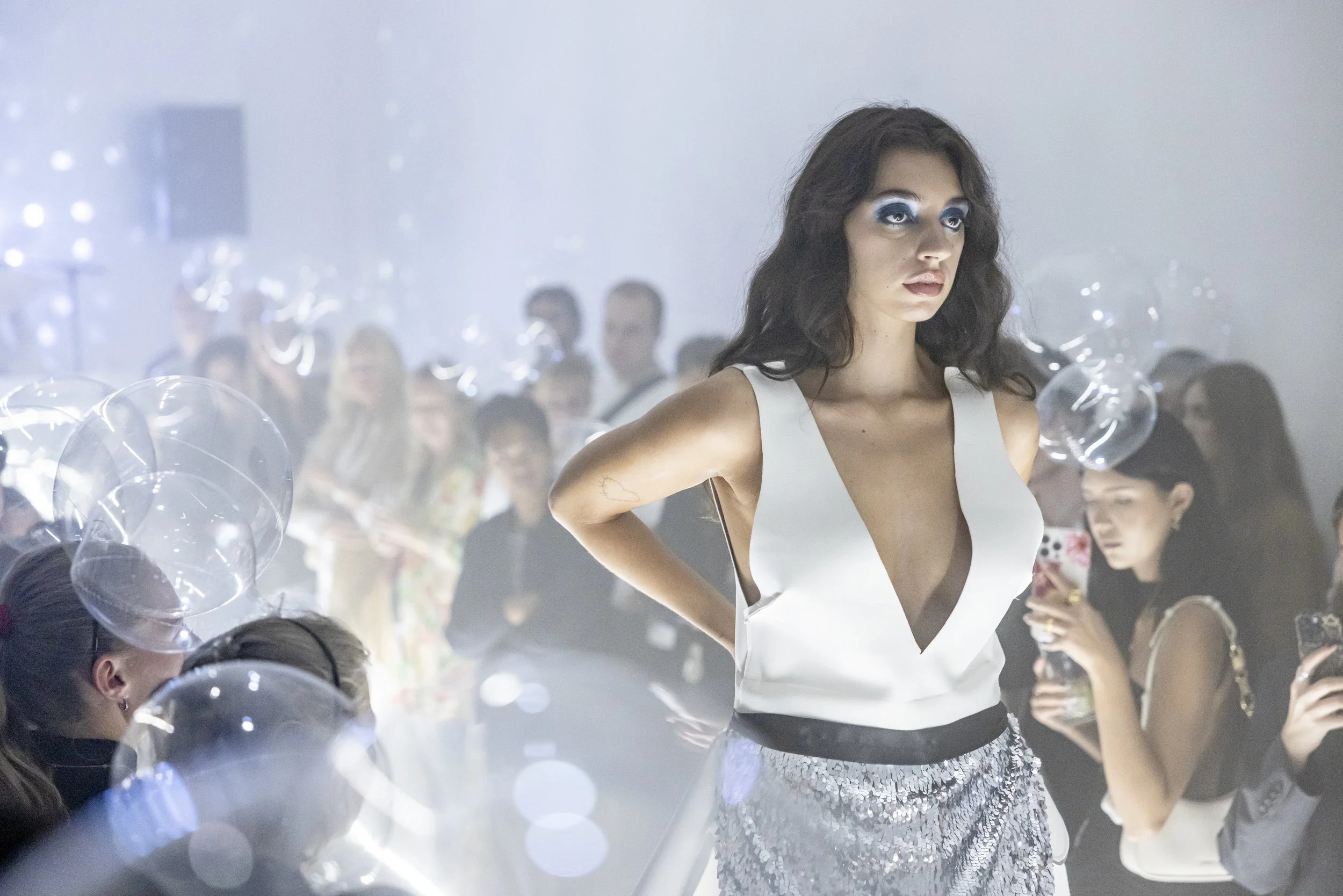 A fashion model walking on a runway at a fashion show with audience members taking photos and balloons in the background.