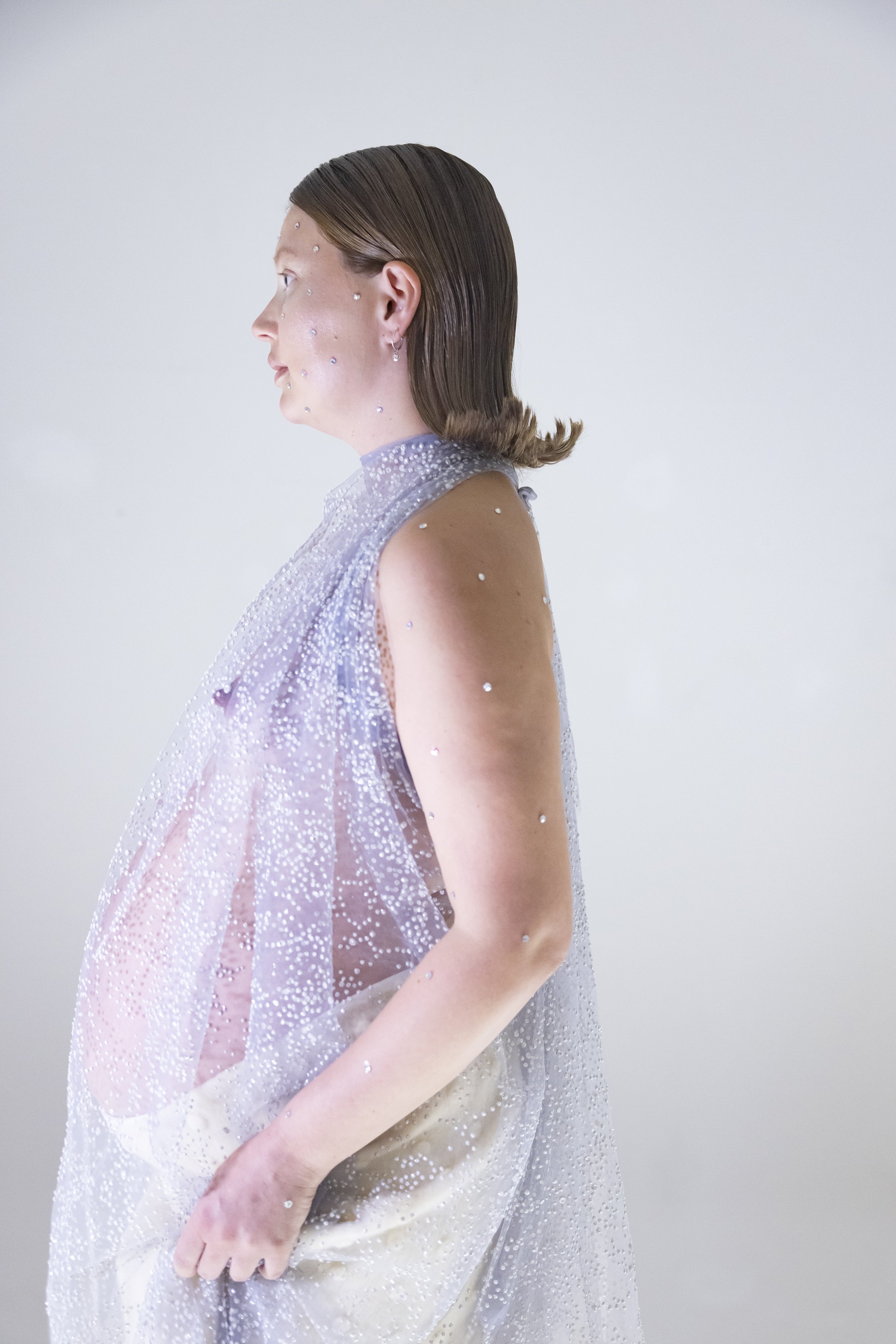 A side profile of a woman with short brown hair, wearing a sleeveless, translucent, sparkly top with droplets and beads, standing against a plain white background.