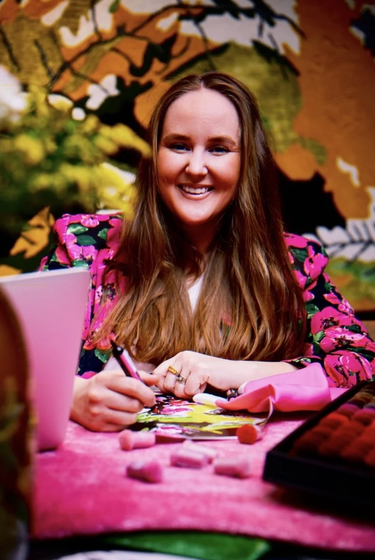 A woman with long brown hair, smiling, sitting at a pink table, signing an item with a pen, surrounded by pink makeup sponges and a pink cup, with a colorful, leafy background.