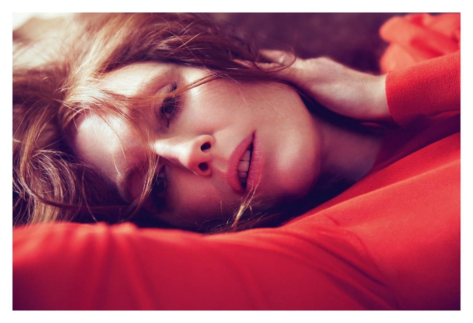 Close-up of a woman with red hair lying down with her eyes closed, wearing a red top, with her hand on her face.