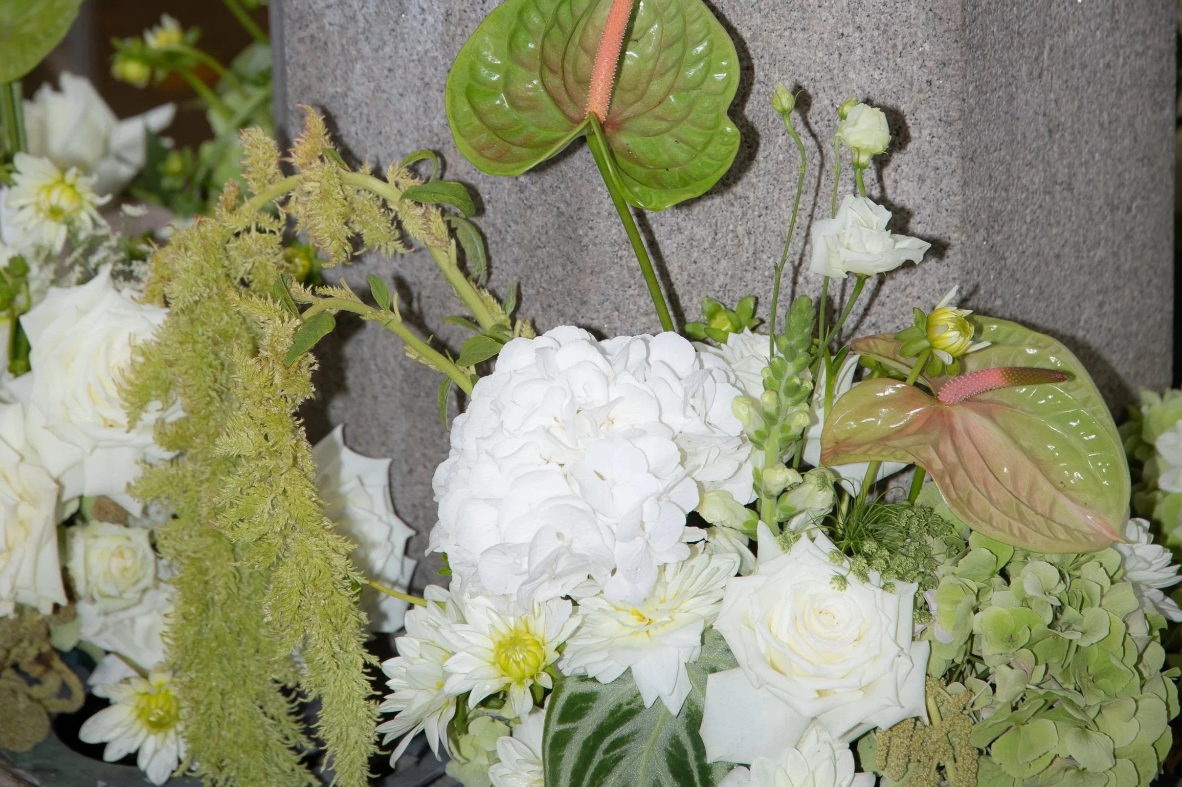 A bouquet of white flowers, including roses and hydrangeas, with green and pink anthuriums and greenery, against a gray stone background.