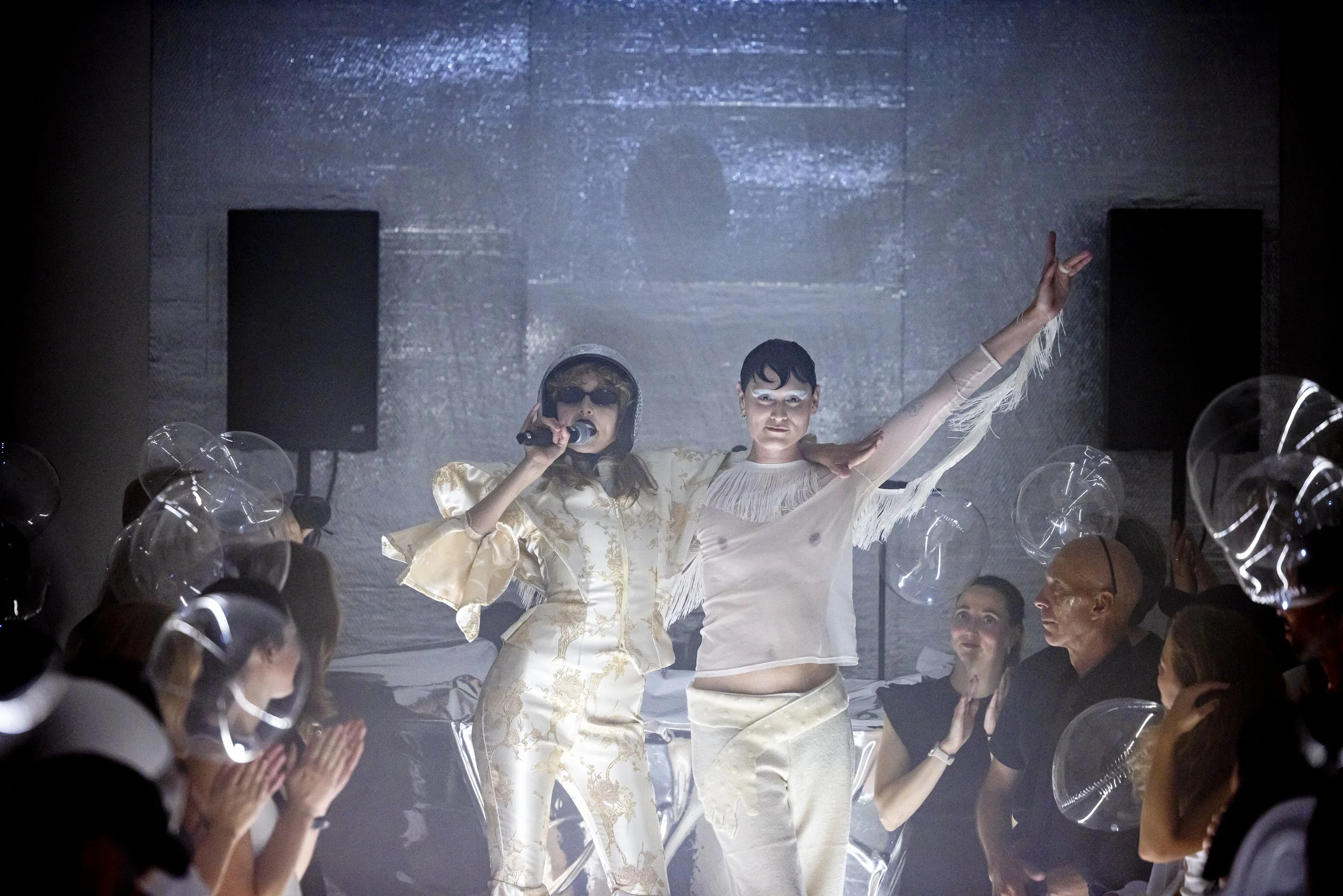 Two women on a stage, one with a microphone and sunglasses, the other with white makeup and a fringed top, performing in front of an audience with holographic balloons.