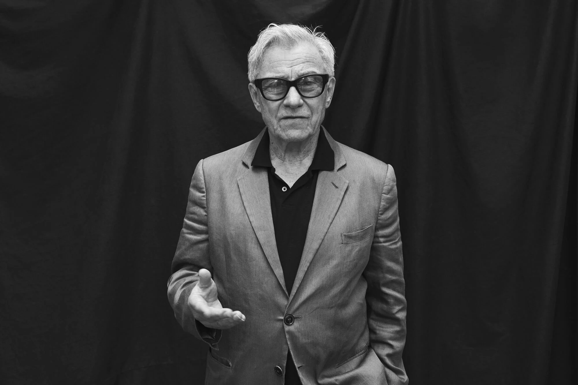 Older man with gray hair, glasses, and a beard, wearing a suit jacket and shirt, standing against a dark curtain background.