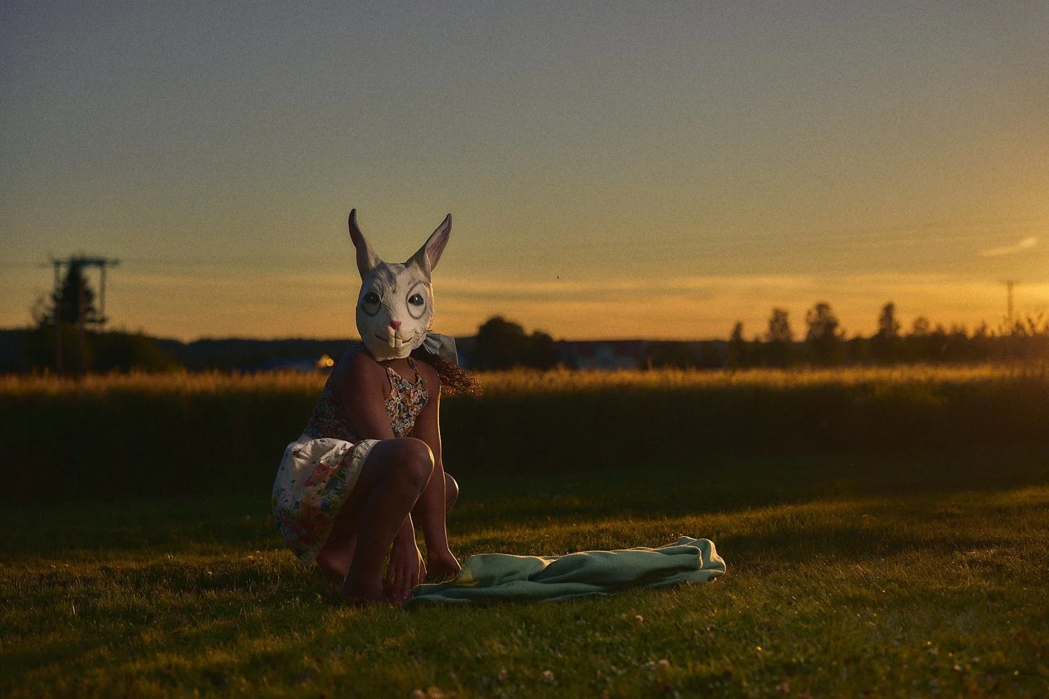 A person wearing a rabbit mask squatting on grass during sunset.