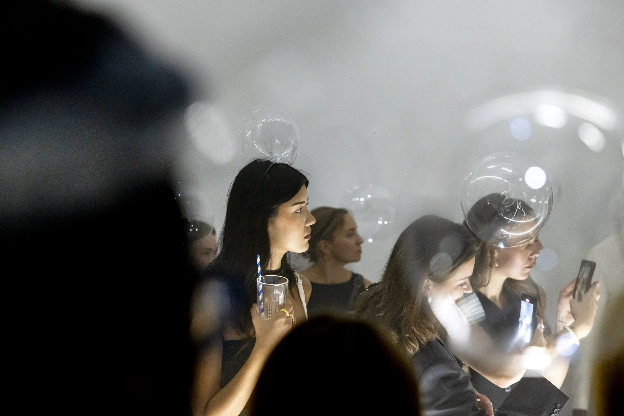 Young women sitting in a row, some looking at their phones, with transparent balloons floating above their heads, in a dimly lit setting.