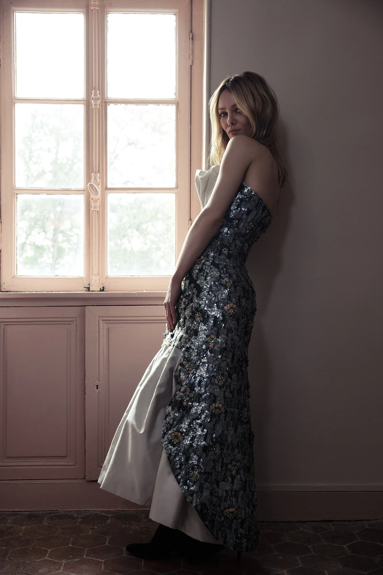 A woman standing inside a room near a window, wearing a strapless, sequined dress with floral patterns and beige fabric accents.