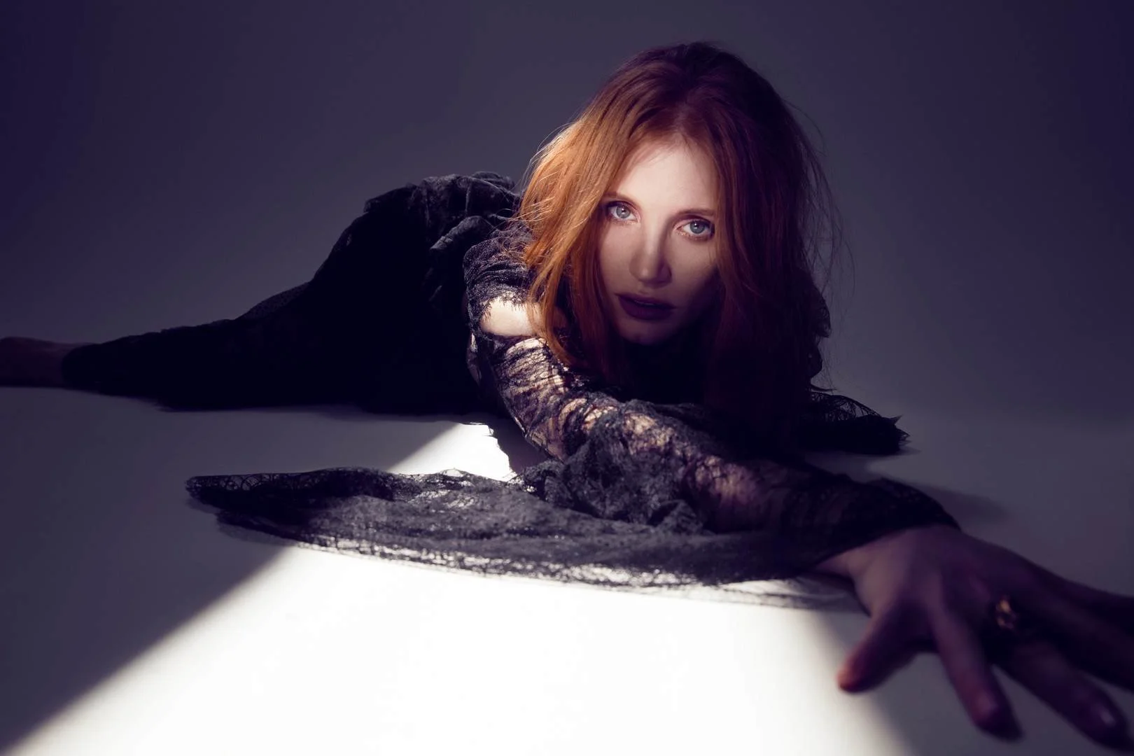 A woman with red hair and fair skin lying on the floor with outstretched arms, wearing black lace clothing, looking directly at the camera with a serious expression, with a shadow cast on the floor.