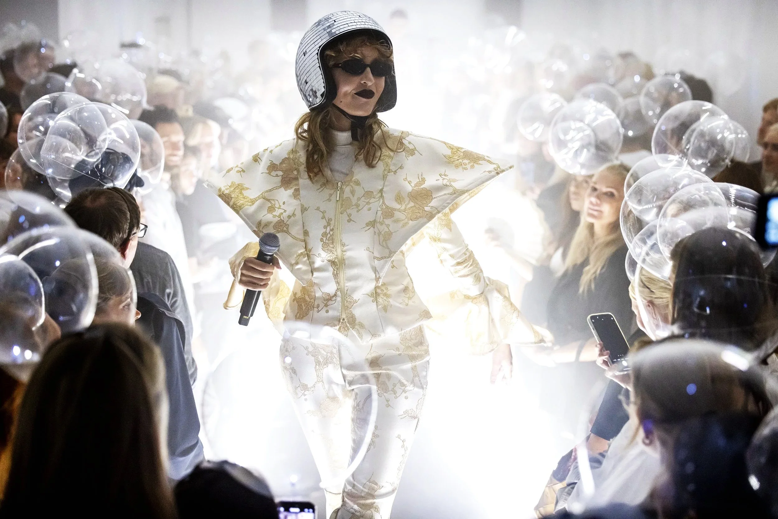 Person dressed in a futuristic, white and gold outfit with a helmet and sunglasses walking through a runway surrounded by an audience holding transparent balloons, amidst bright lighting.
