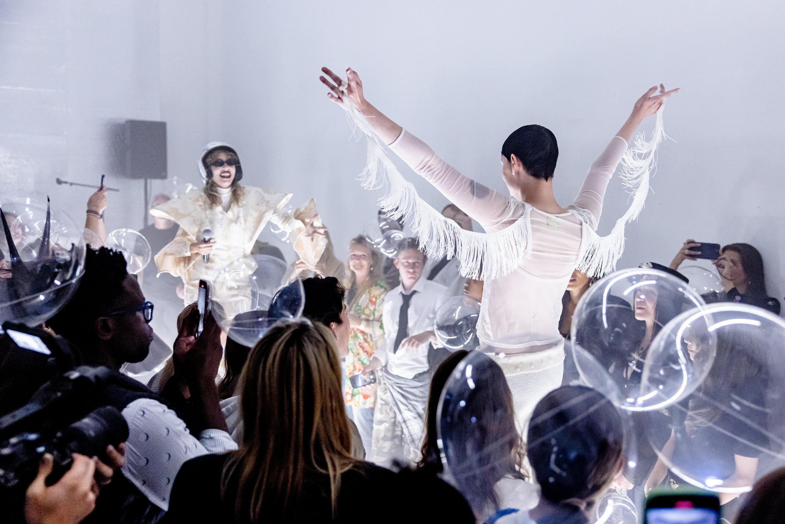 A performer with short black hair in white fringed clothing dancing with arms raised in front of a seated crowd. The crowd features people in dark glasses and some with cameras, with a woman in the background wearing a helmet and sunglasses, holding a microphone among transparent balloons.