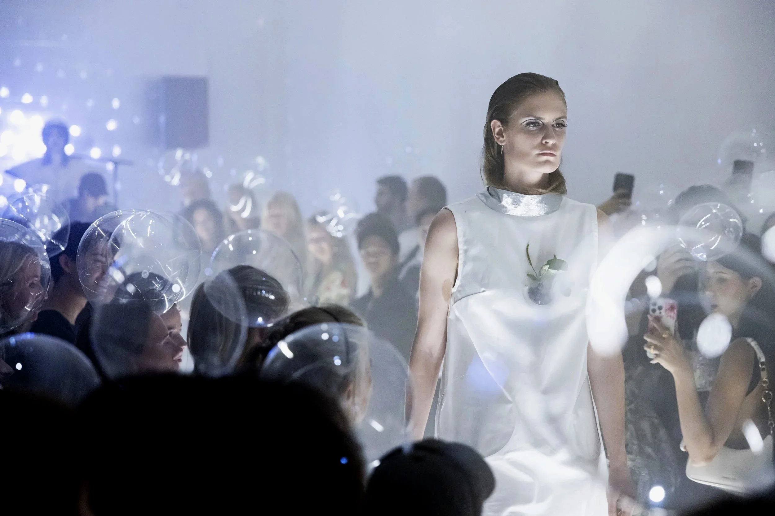 A fashion show runway with a model walking in a white dress with metallic accents, surrounded by an audience taking photos, some with transparent balloons and glowing lights.