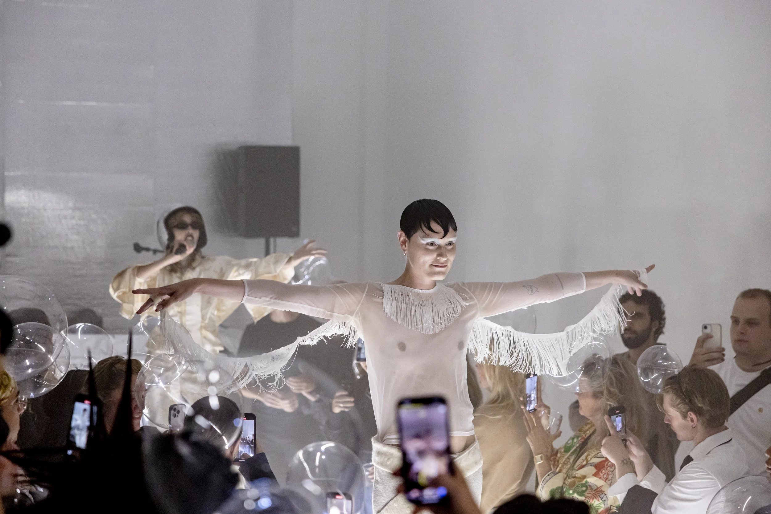 A performer in a sheer, white fringed top dances with arms outstretched on a stage in front of an audience, with some audience members capturing the moment on their phones.