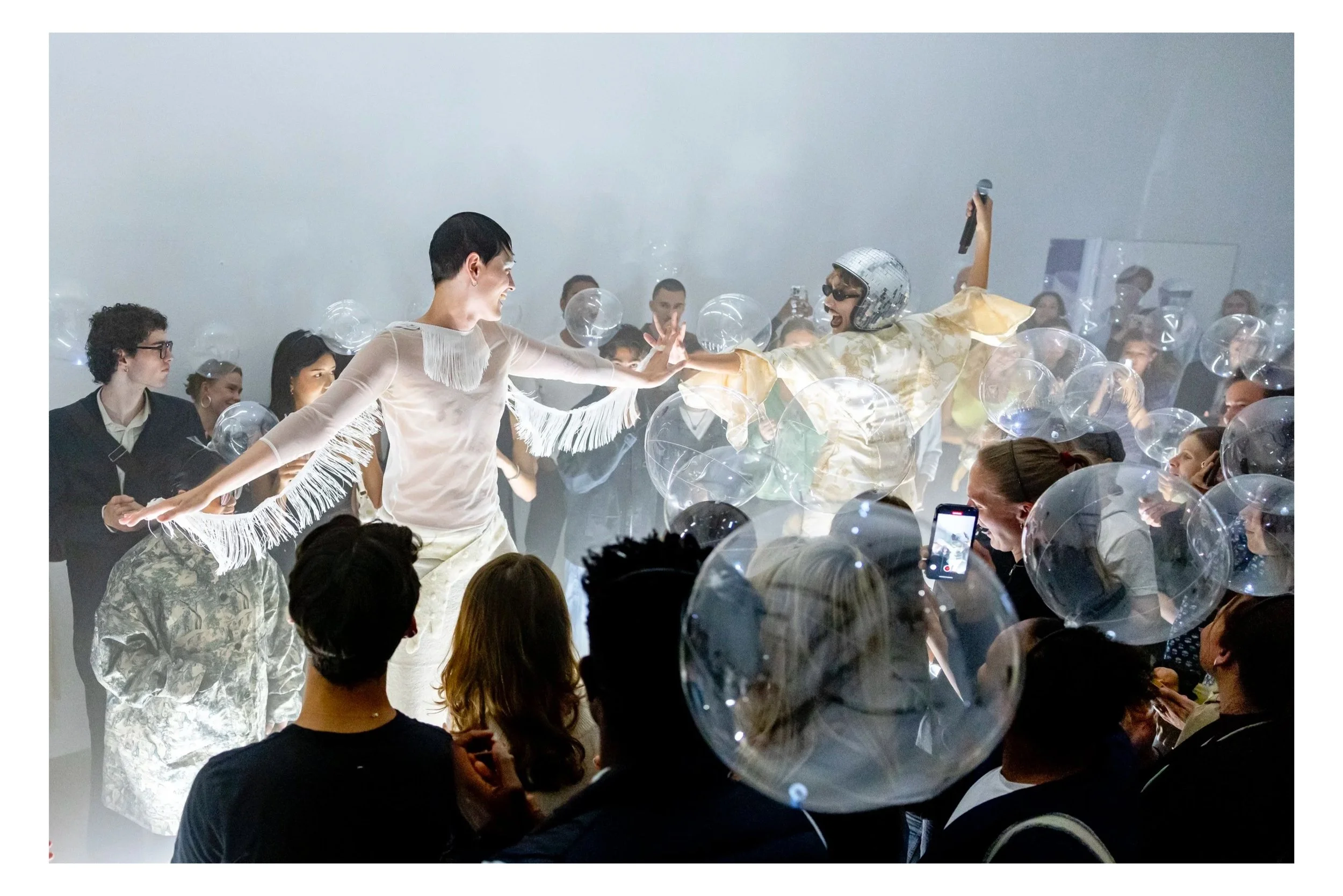 People dancing and enjoying a lively party with balloons and someone singing into a microphone, surrounded by an audience taking photos.