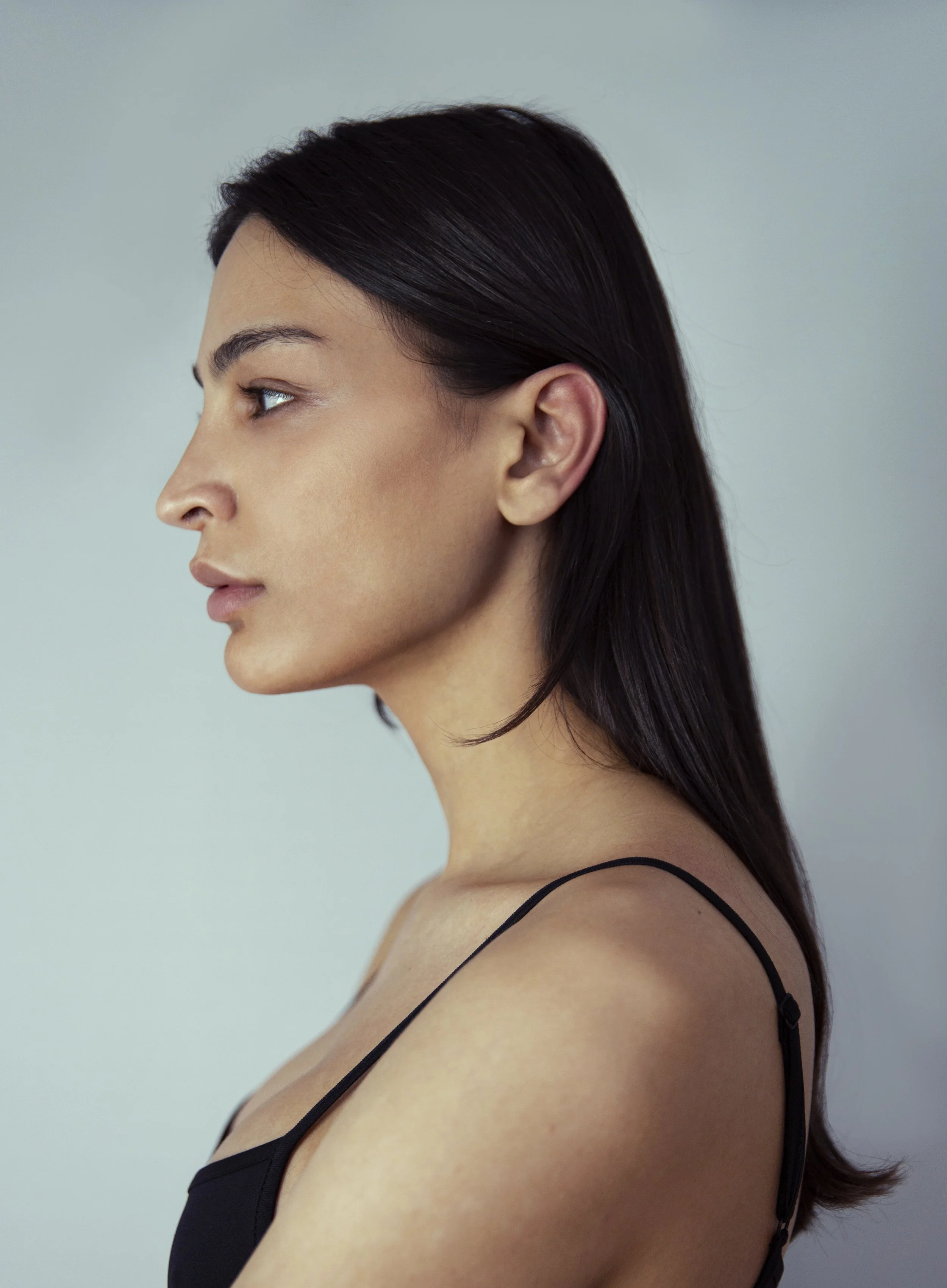 Side profile of a woman with long, dark hair and light skin, wearing a black strap top, against a plain background.