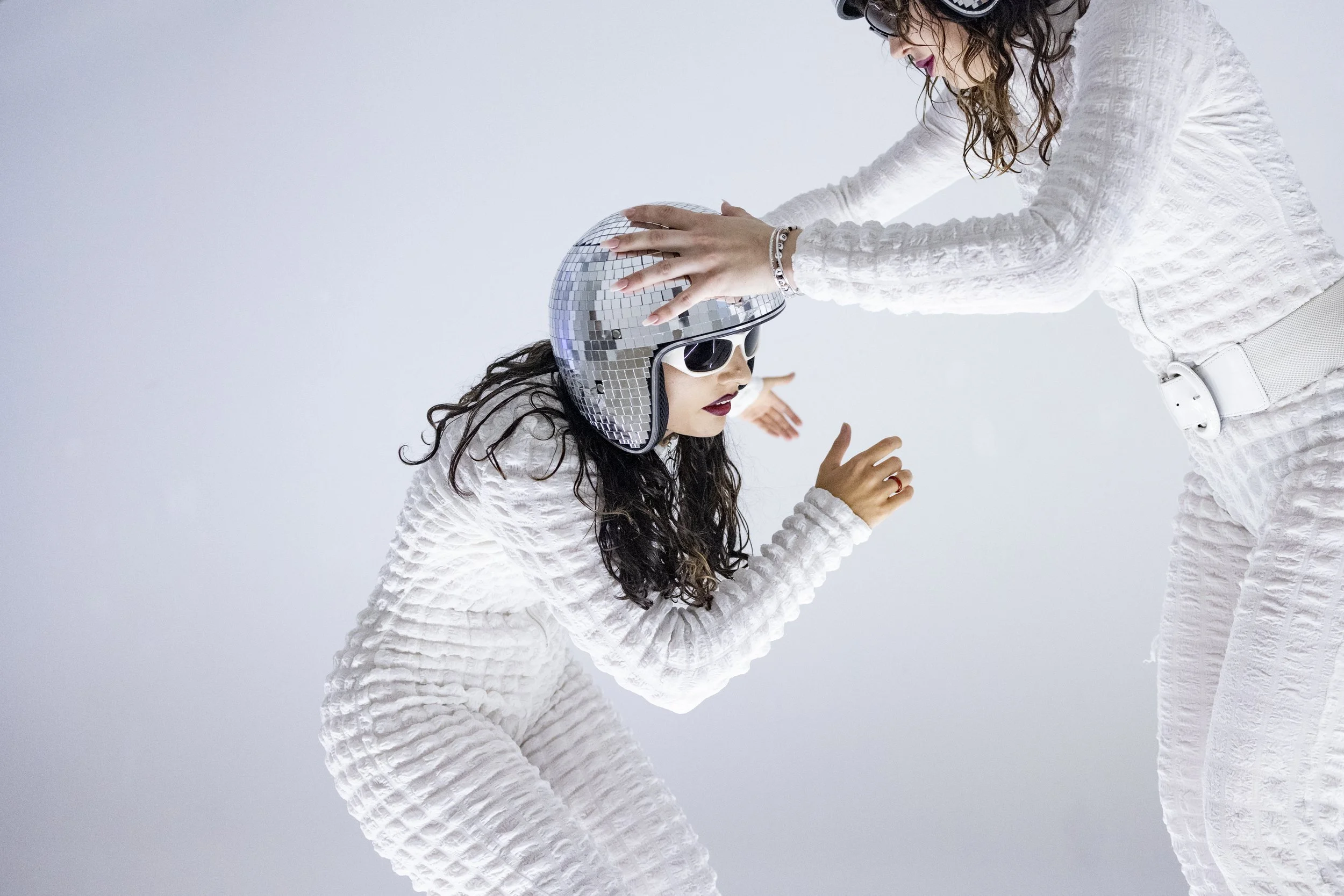 Two women dressed in white, one wearing a reflective disco helmet and sunglasses, in a playful pose against a plain background.