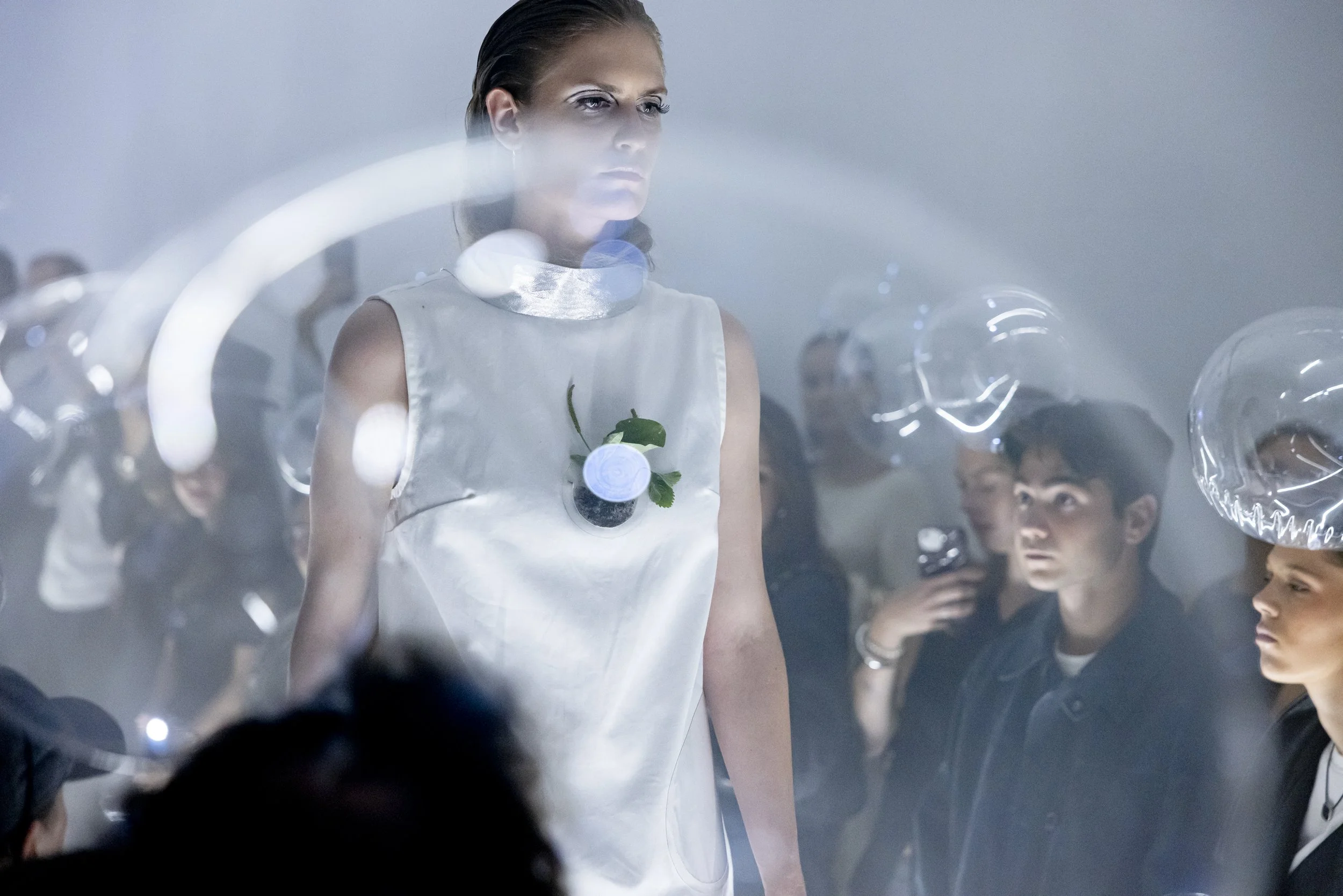A woman with futuristic fashion and makeup standing among people wearing transparent helmets in a smoky, high-tech setting.