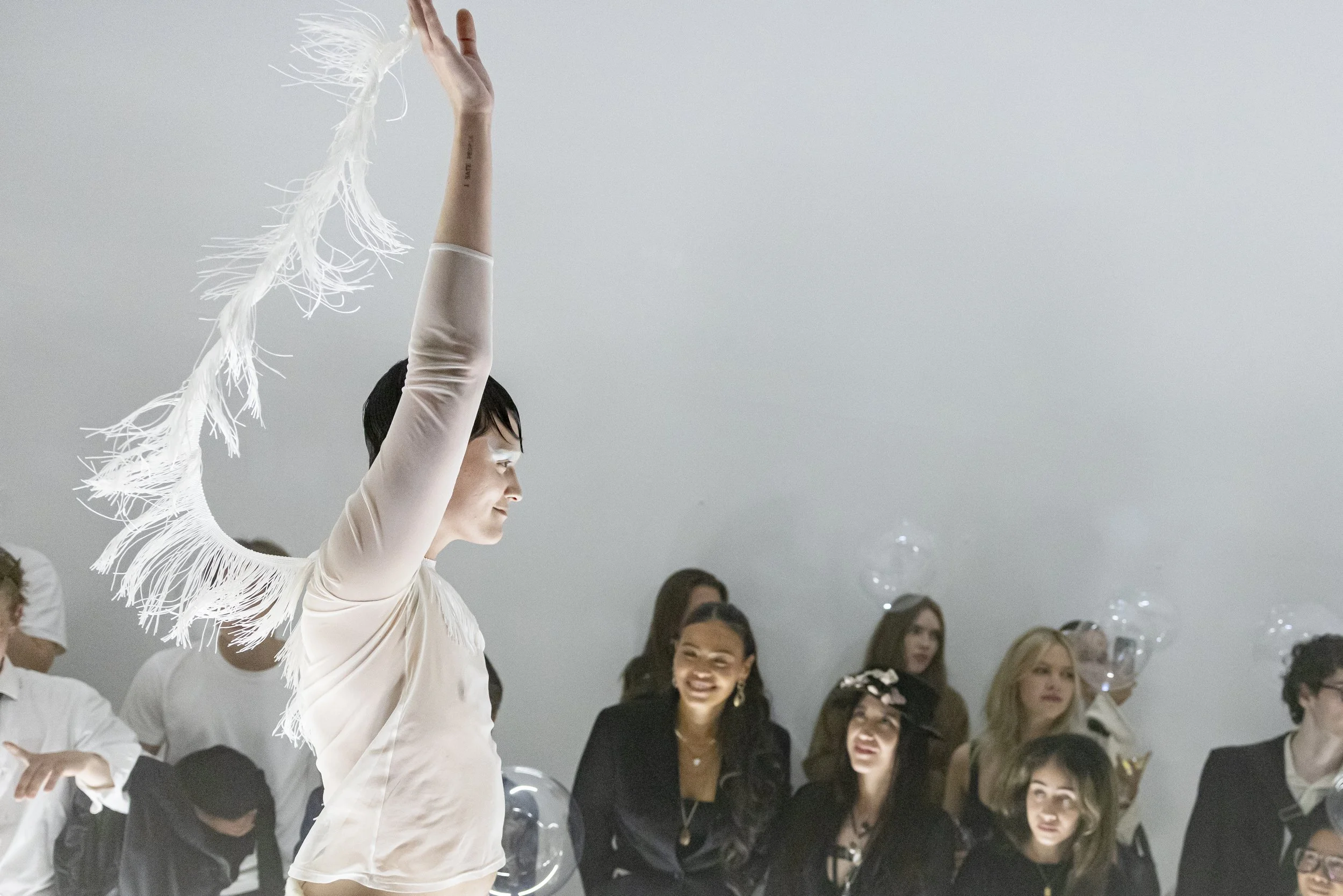 A woman in a light-colored dress with fringes on her sleeves raises one arm while standing in front of an audience of smiling women, some with balloons and transparent bubbles, in a bright, minimal room.