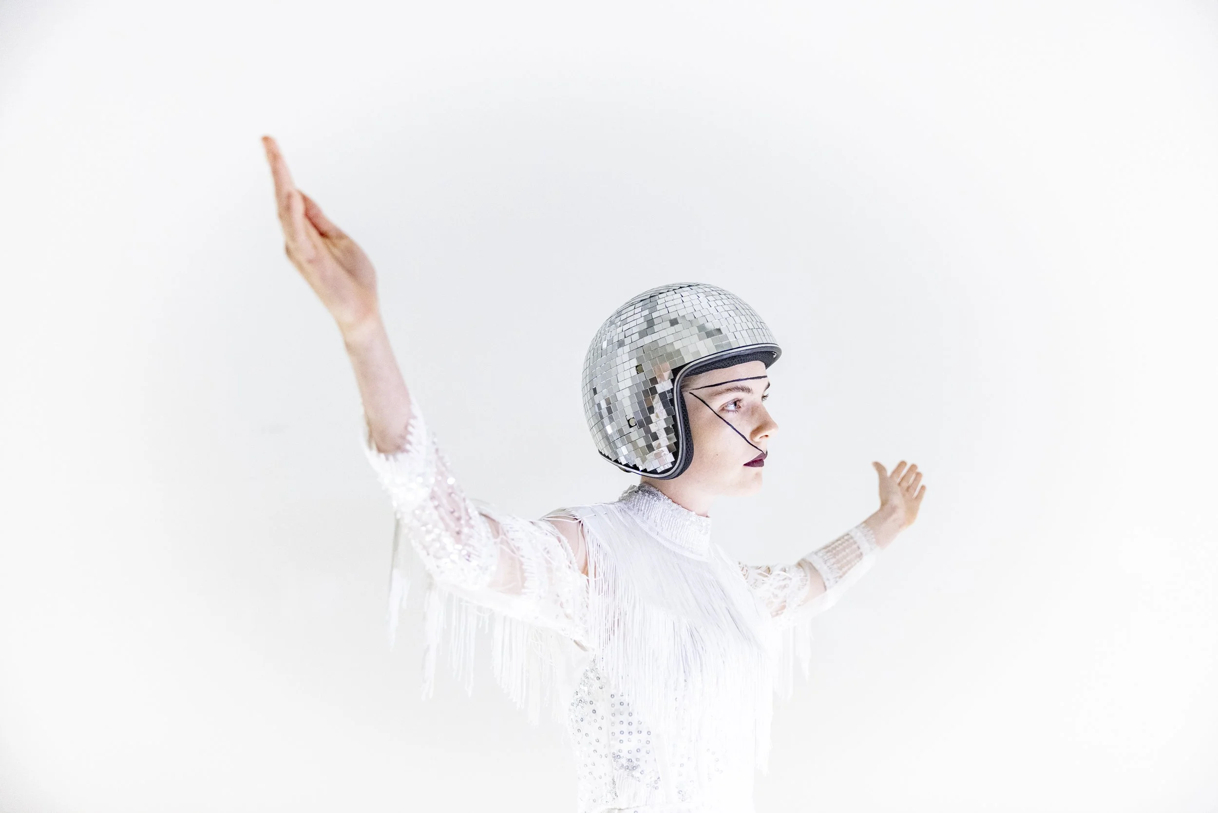 A woman wearing a shiny disco helmet, with arms raised, dressed in a white, fringed outfit, posing against a plain white background.