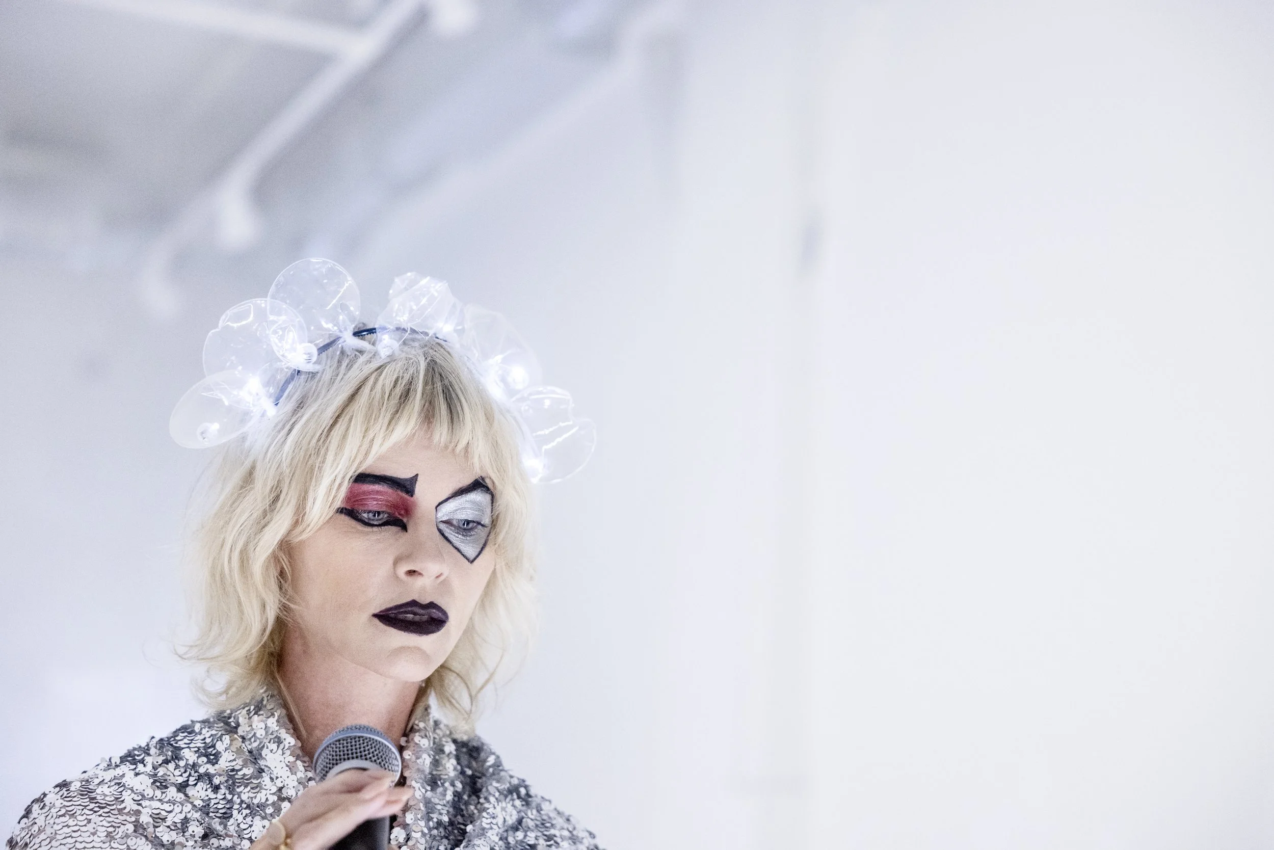 Woman wearing bold, colorful makeup with a white, floral headpiece, holding a microphone, against a plain, white background.