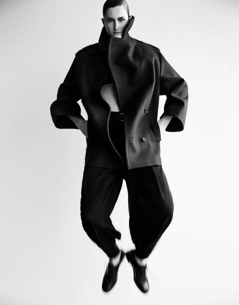 Black and white fashion photograph of a woman in a stylish black coat with oversized sleeves, baggy black pants, and black shoes, standing against a plain light background.