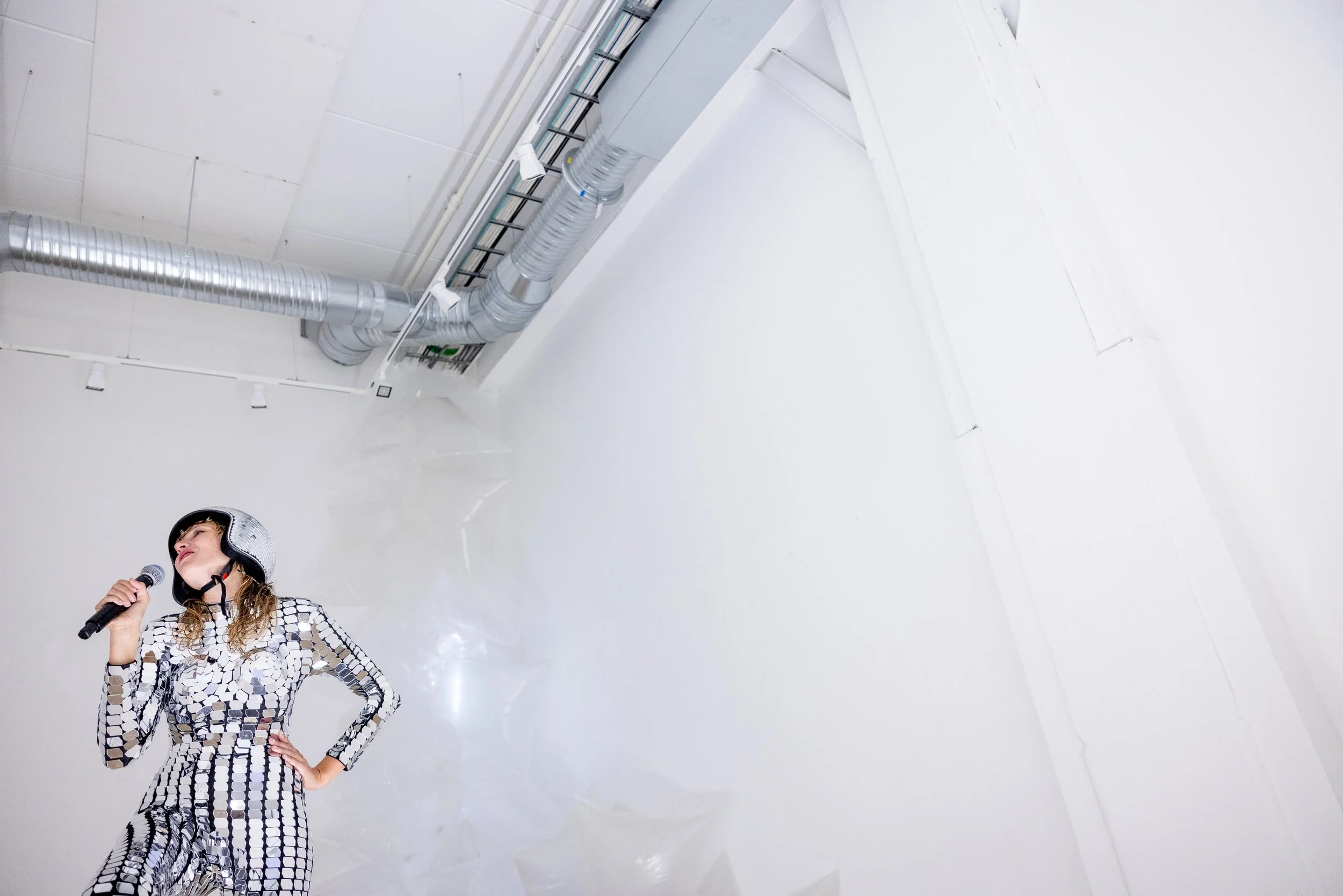 A woman wearing a matching black and silver geometric patterned outfit and a helmet, holding a microphone, standing in a white room with exposed industrial pipes on the ceiling.