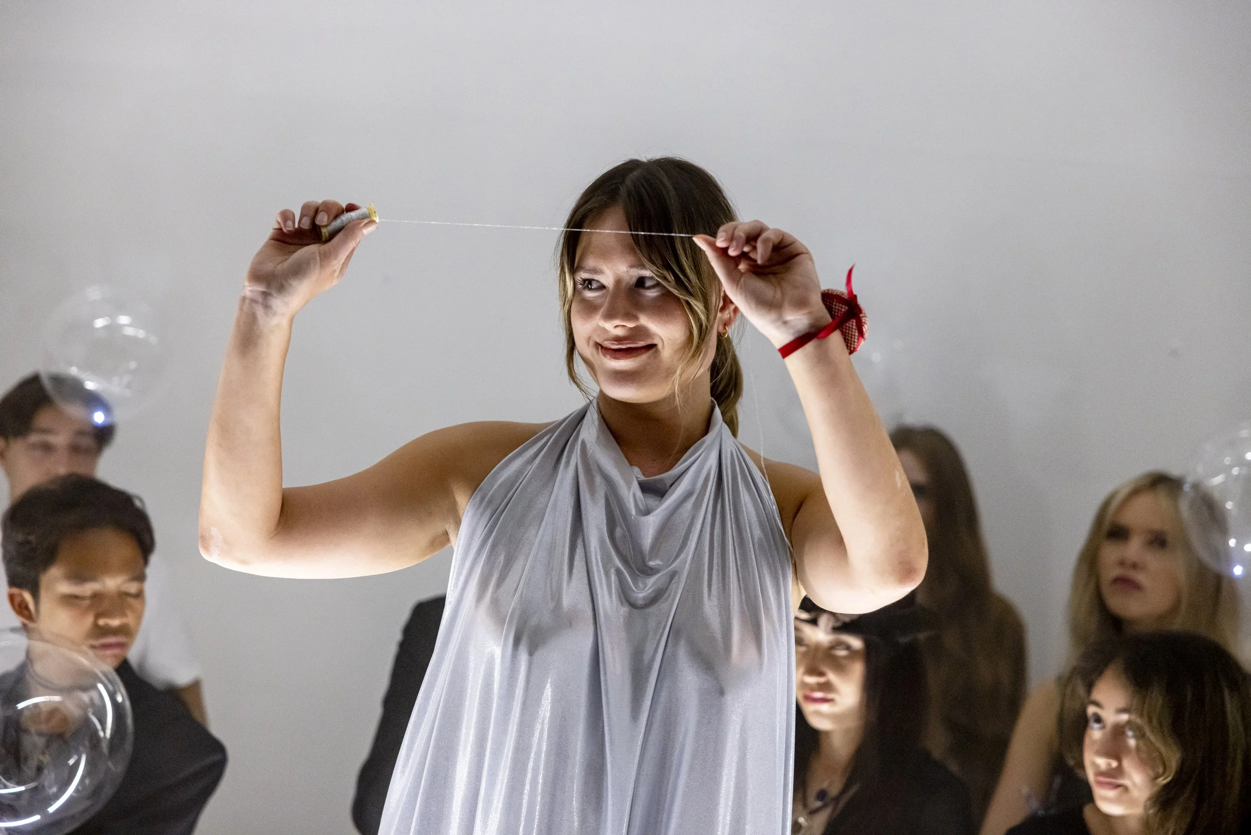 A woman is making a bubble with a string tool at a party, with several onlookers in the background.