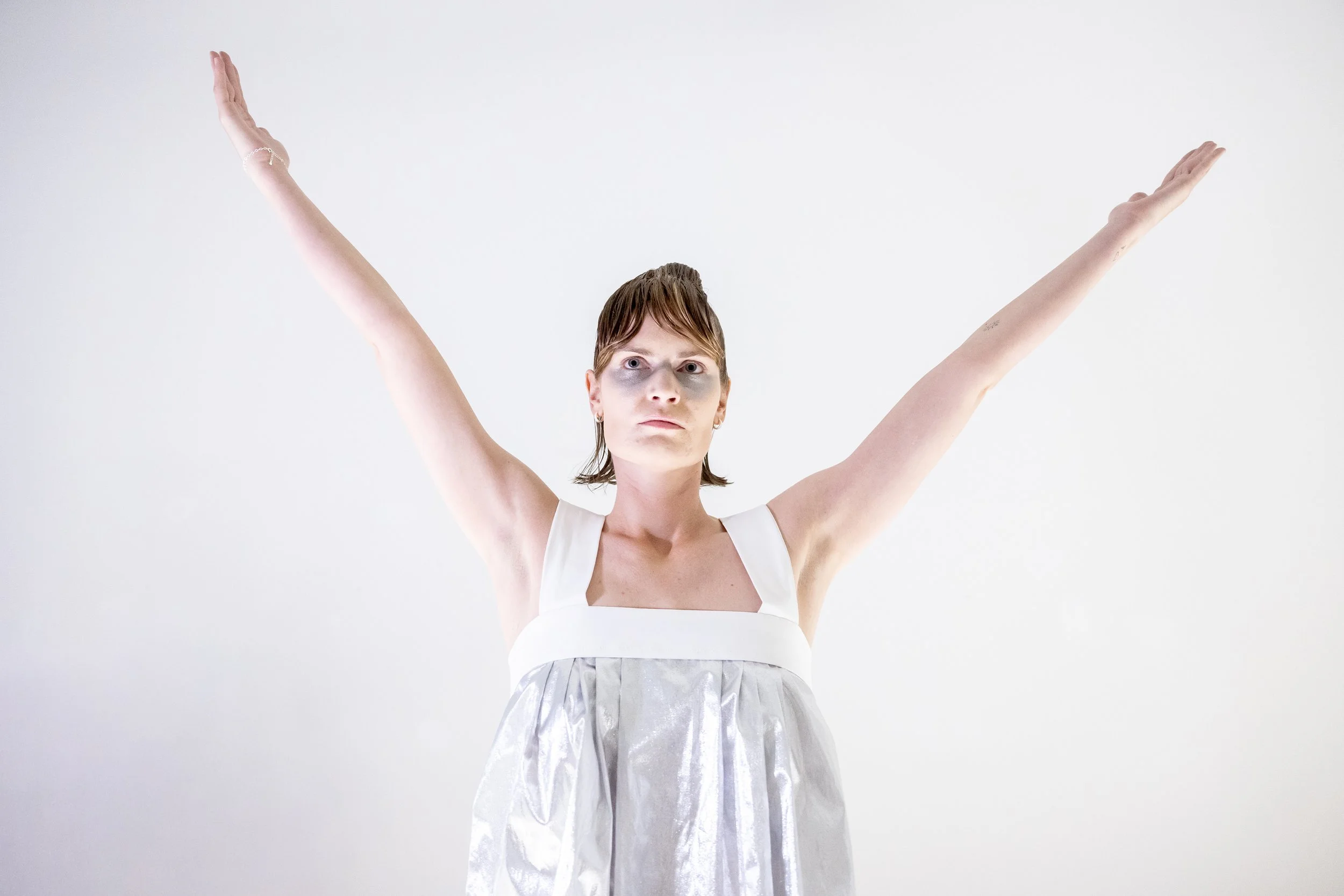 Person with short brown hair standing against a plain white background, wearing a sleeveless silver and white dress, with arms raised and looking directly at the camera.