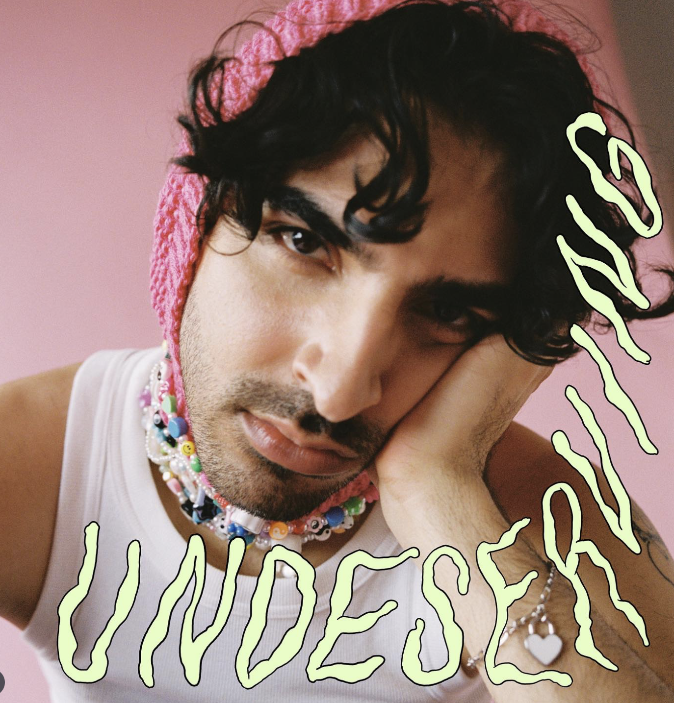 A person with black curly hair, wearing a pink knit hat and colorful beaded necklace, resting their head on their hand against a pink background, with the words 'SAD UNDESE' written in light green on the image.