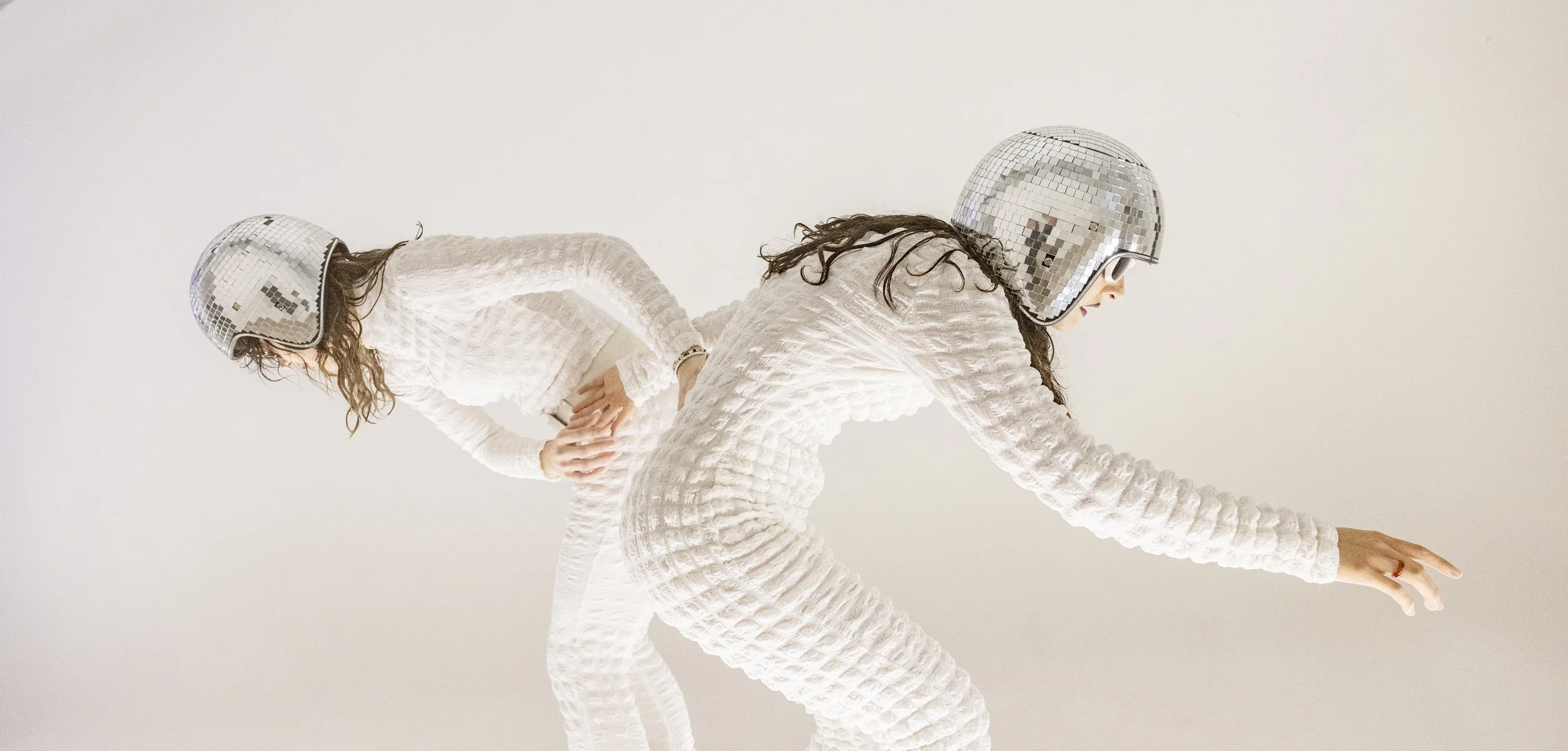 Two women wearing textured white outfits and metallic disco helmets, with the woman in the foreground reaching forward and the woman in the background bending backward.