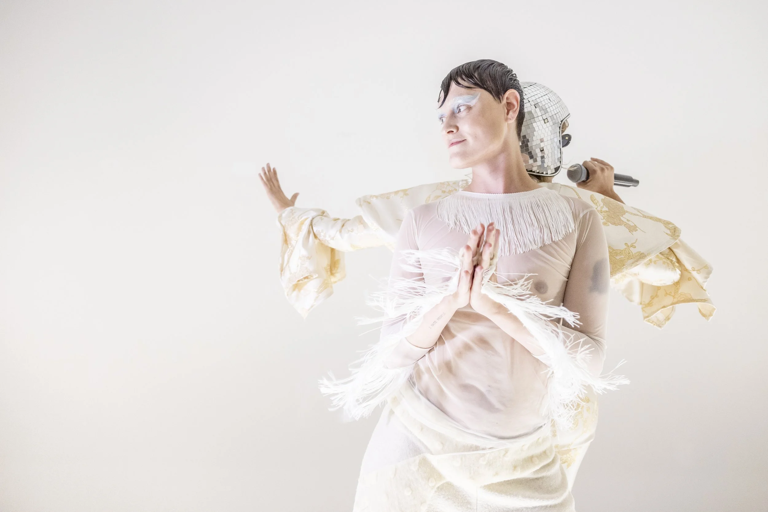 A person wearing a sheer, fringed top and white fabric with gold patterns, posing with hands pressed together, standing in front of a white background, with a performer wearing a shiny disco ball helmet and holding a microphone behind them.