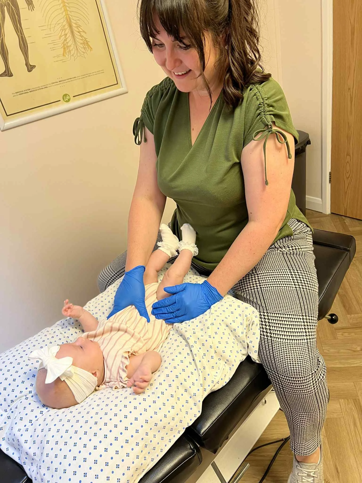 A woman with brown hair, wearing a green top and checkered pants, is examining a baby on an examination table at a doctor's office.