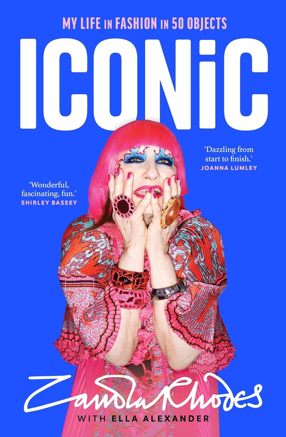 Book cover with colorful design featuring person with vibrant hair and bold makeup, wearing ornate jewelry. Text reads "Iconic: My Life in Fashion in 50 Objects", with quotes from Shirley Bassey and Joanna Lumley. Authors: Zandra Rhodes with Ella Ale