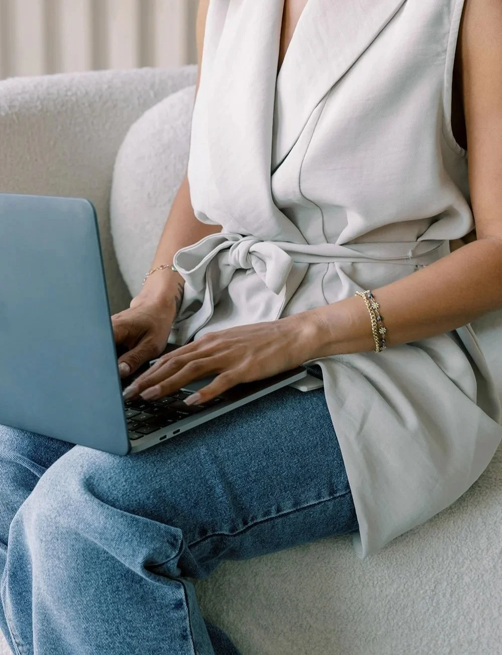 A woman in a sleeveless beige top and blue jeans sitting on a couch, typing on a silver laptop.