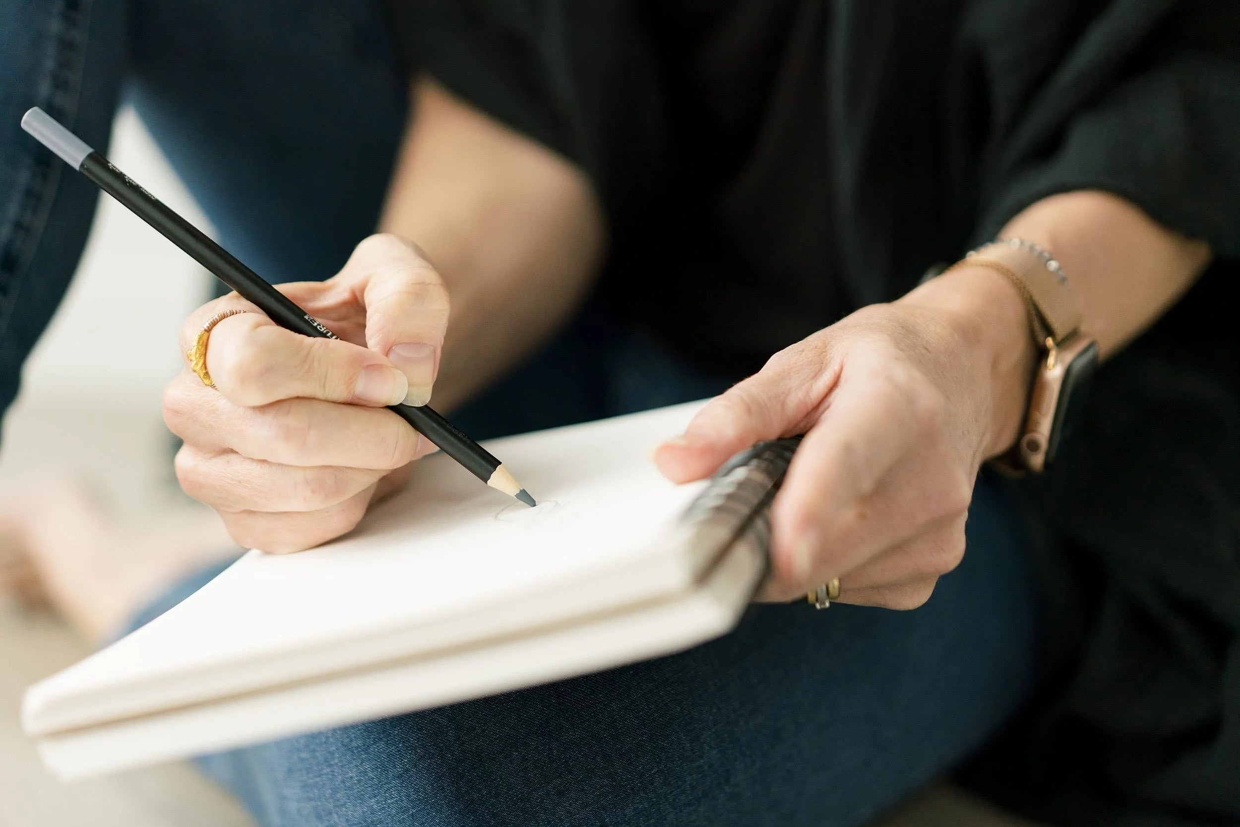 Close-up of an interior designer sketching in a notepad with a black pencil, wearing rings, bracelets, and a watch, sitting on a textured dark blue surface.