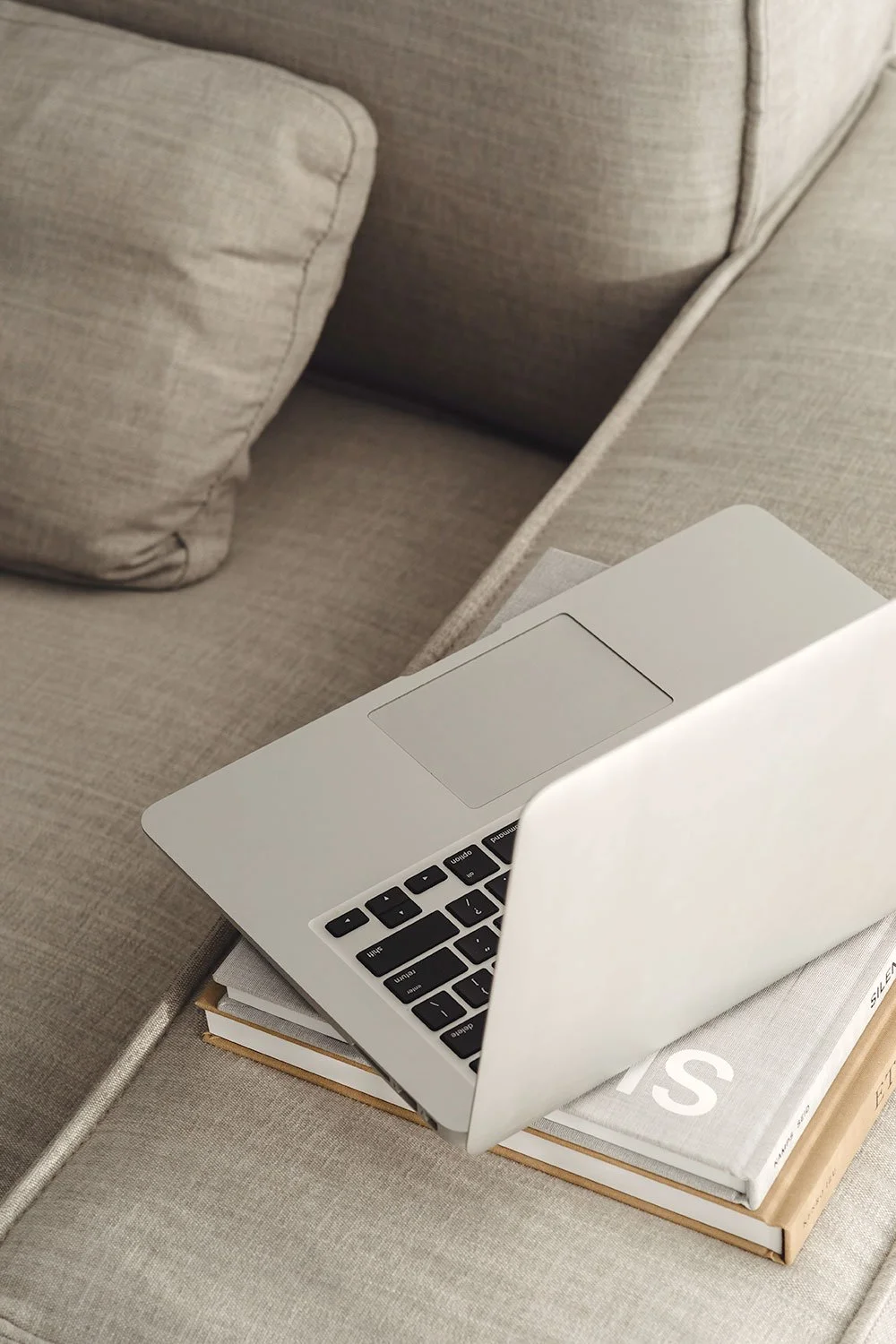 A silver laptop open on top of two books, placed on a beige sofa with a cushion.