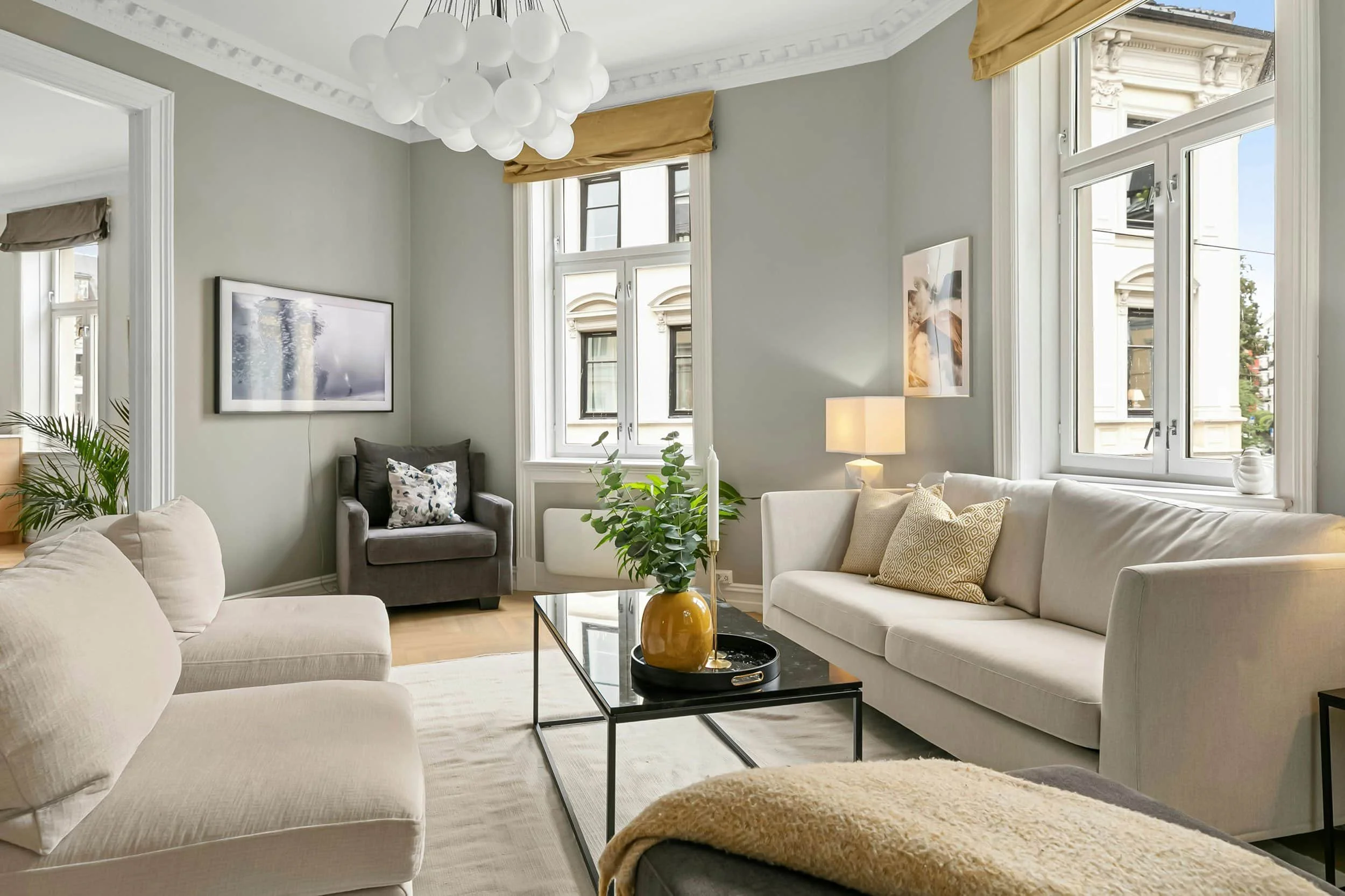 A contemporary living room with large windows, cream sofas, a gray armchair, a black coffee table with a yellow vase and green plant, and minimalist decor including framed artwork, lamp, and throw pillows.