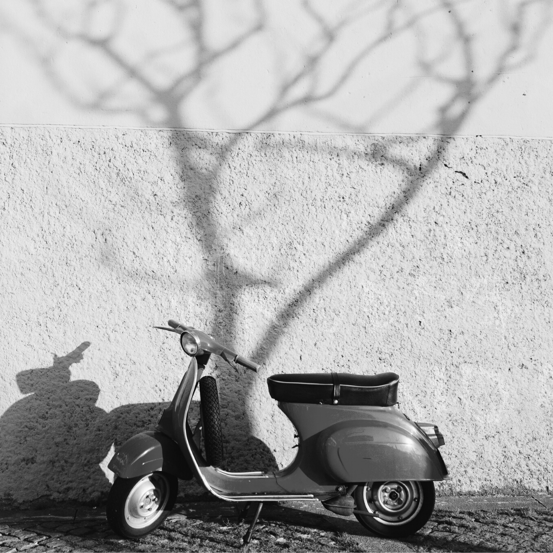 A vintage scooter parked against a textured wall, with a shadow of a tree cast on the wall in black and white.
