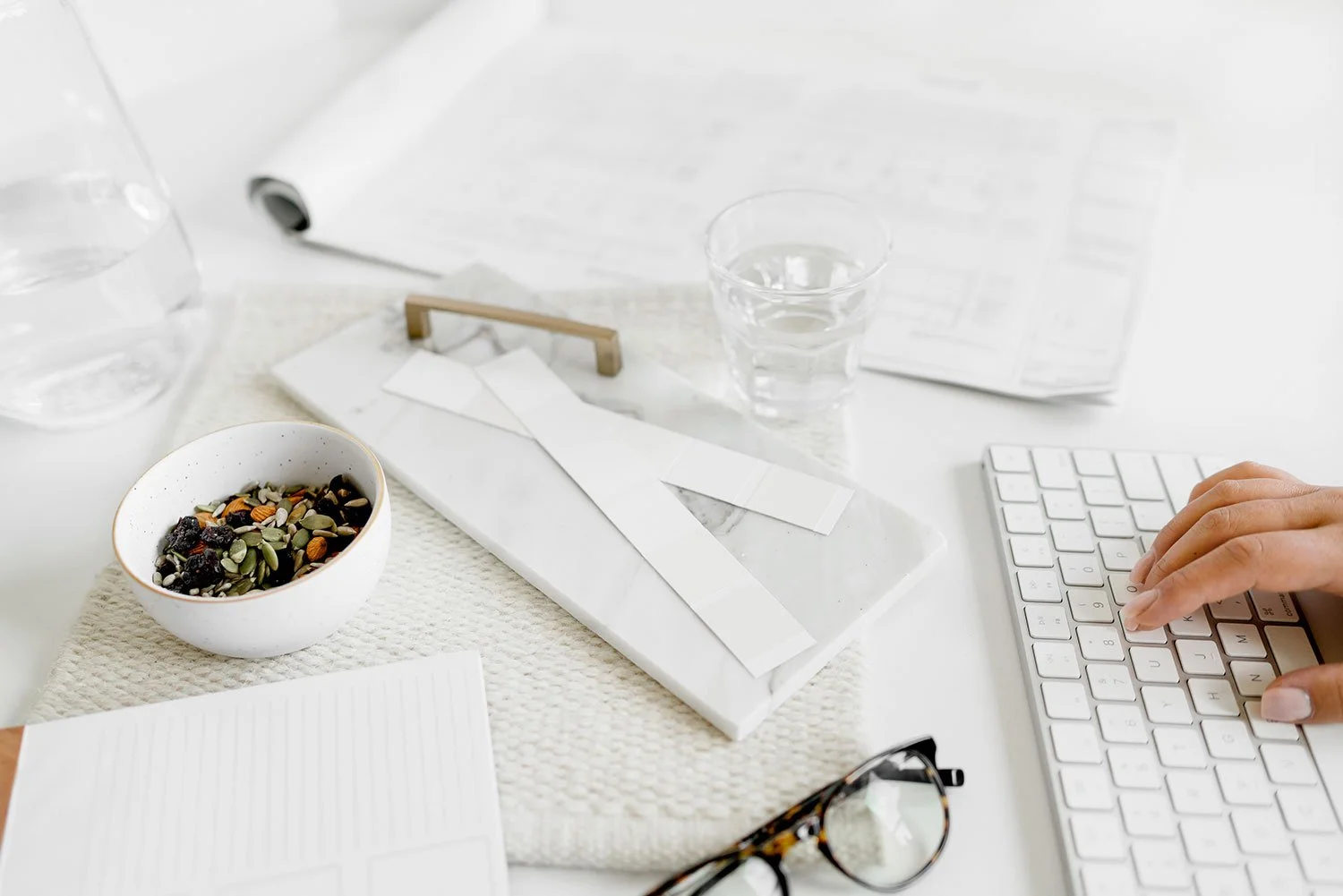 Desktop workspace with a person's hand typing on a white keyboard, a glass of water, a bowl of trail mix, a pair of glasses, a notebook, some scattered papers, and a glass of water, all on a white surface.