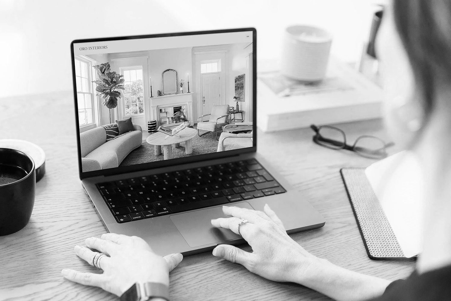 Person working on a laptop at a desk, viewing a living room interior on the screen, with a cup of coffee, glasses, and a notebook on the desk.