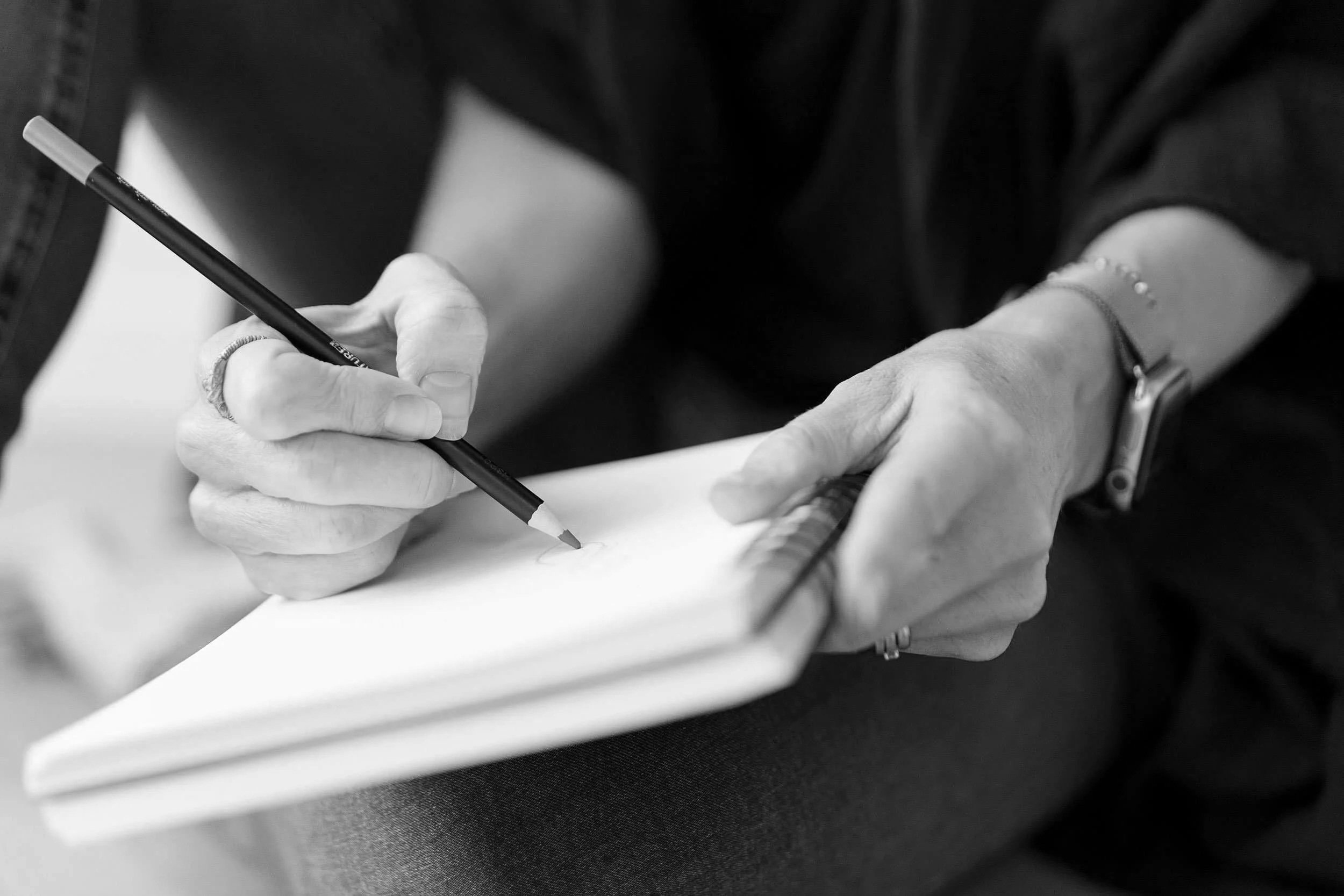 Black and white photo of a person writing with a pen on paper, wearing a bracelet and watch, with a ruler visible nearby.