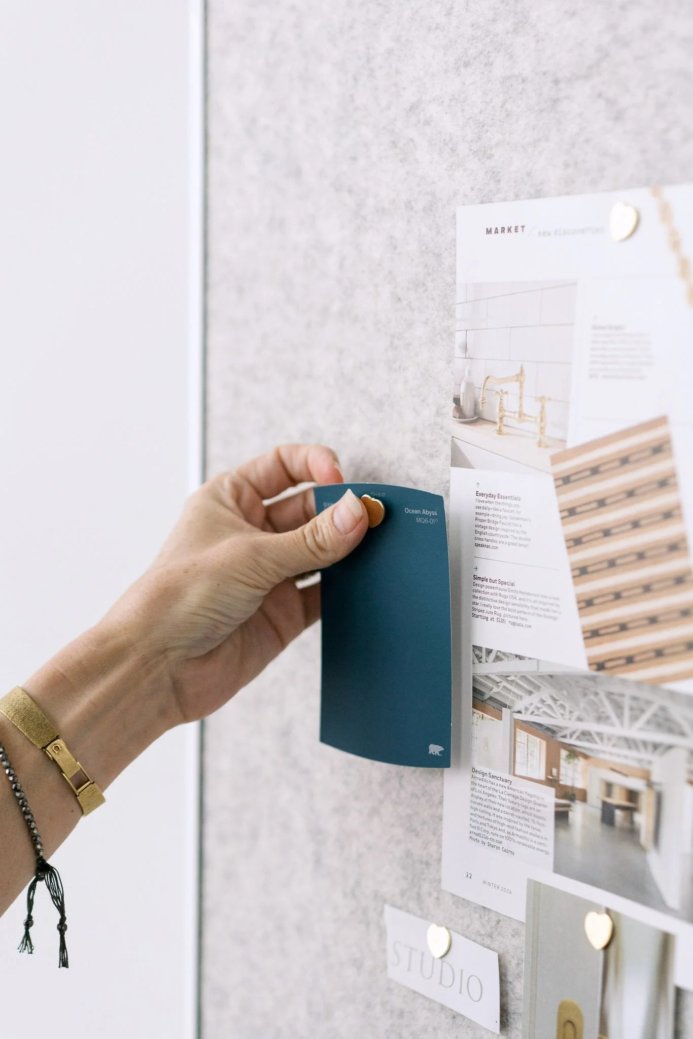 Hand with a gold bracelet pinning a blue card to a grey bulletin board with various papers and photos.